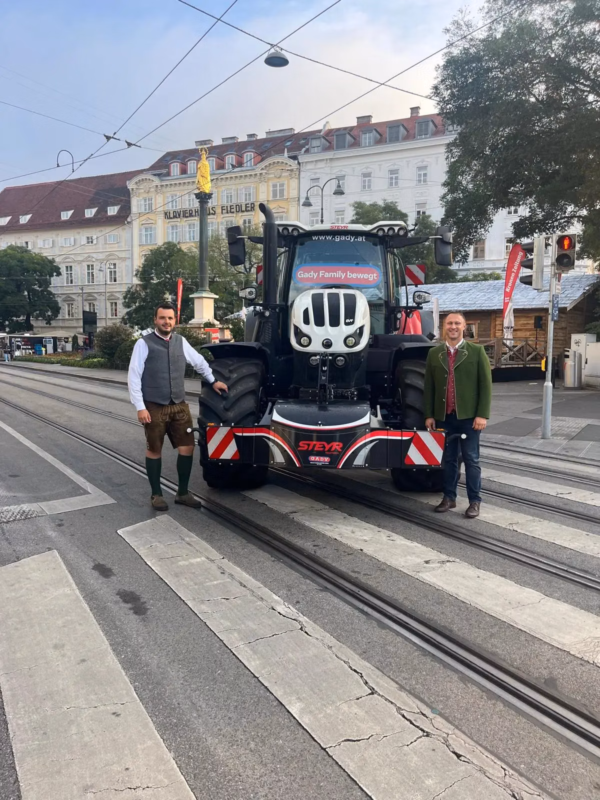 Two men stand in front of a tractor equipped with a Steyr front weight bumper on a city street crosswalk