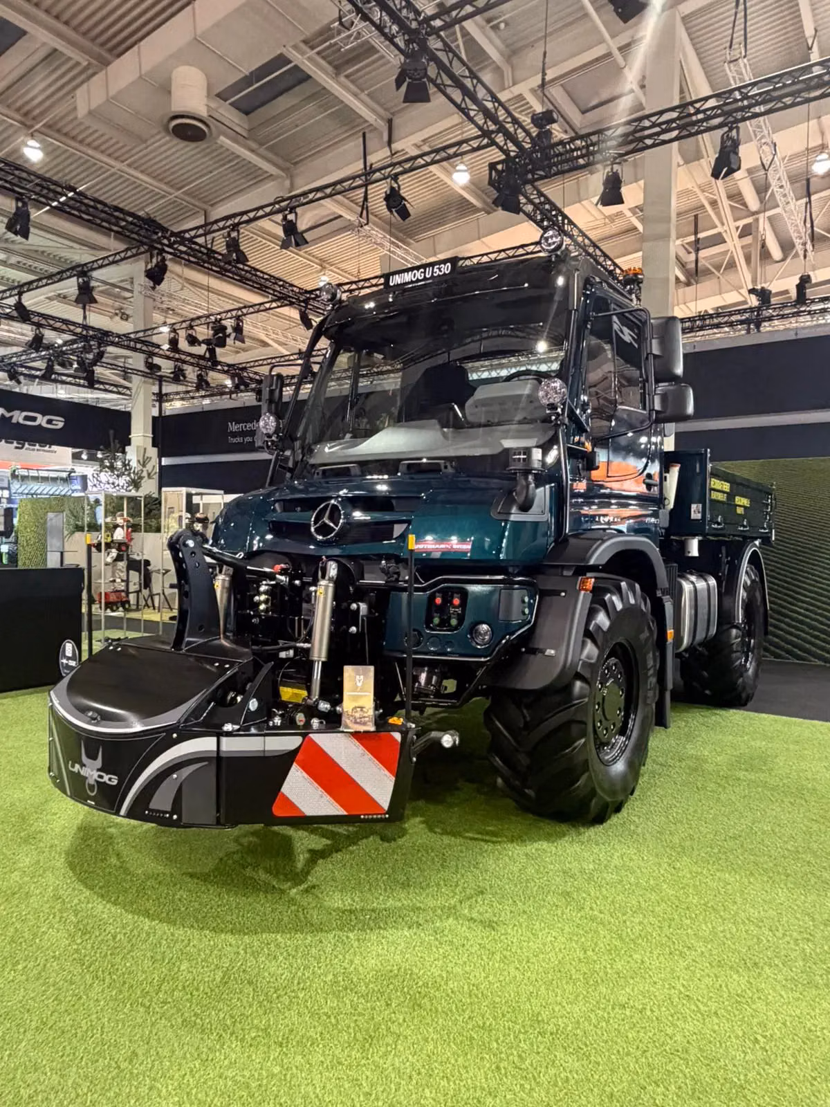 A large dark green utility vehicle with a front-mounted weight bumper featuring red and white reflective stripes