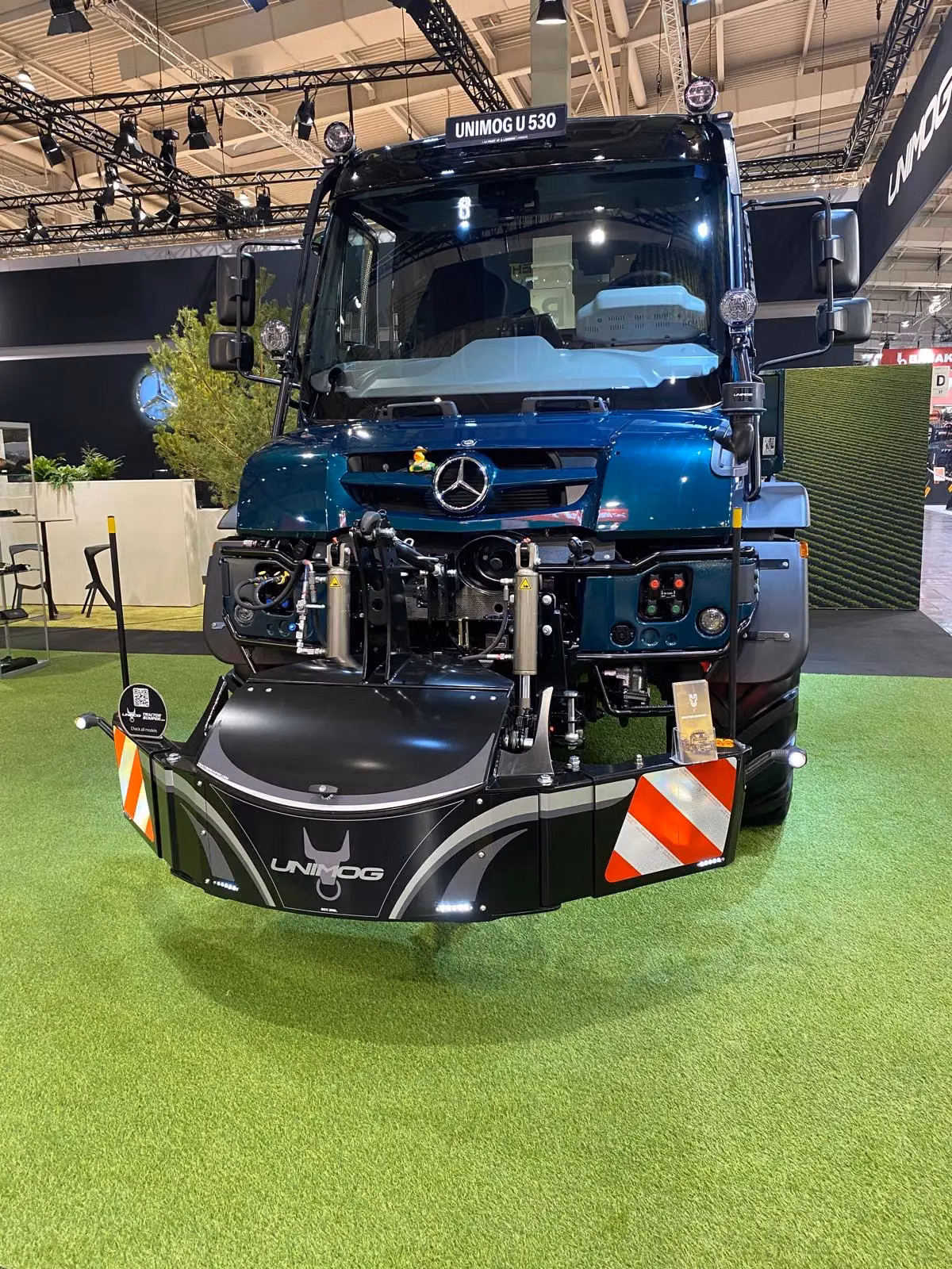 A Unimog vehicle is displayed indoors with a black front weight bumper featuring reflective red and white stripes