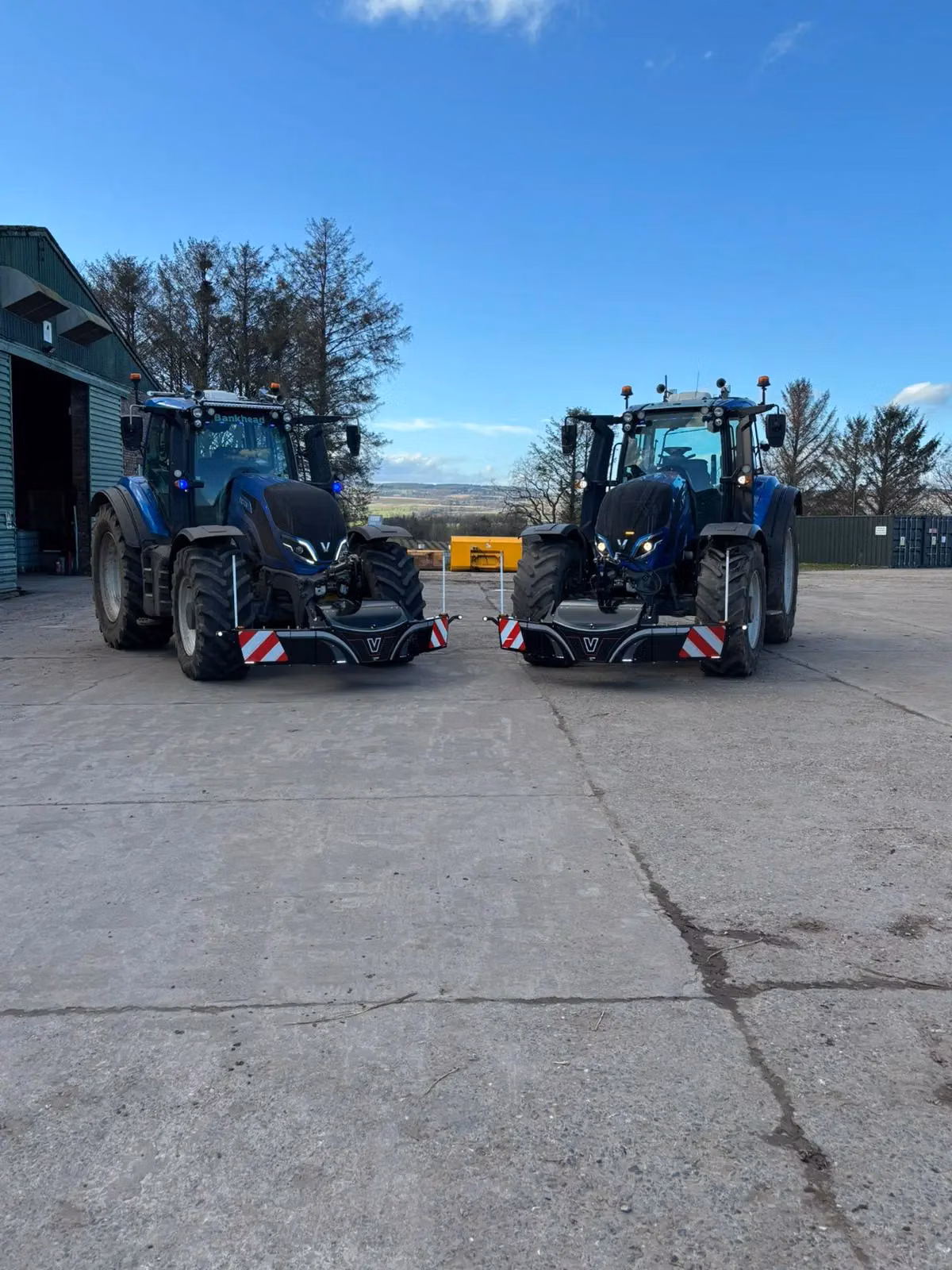 Two blue tractors fitted with black front weight bumpers featuring LED lights, storage boxes and safety markings