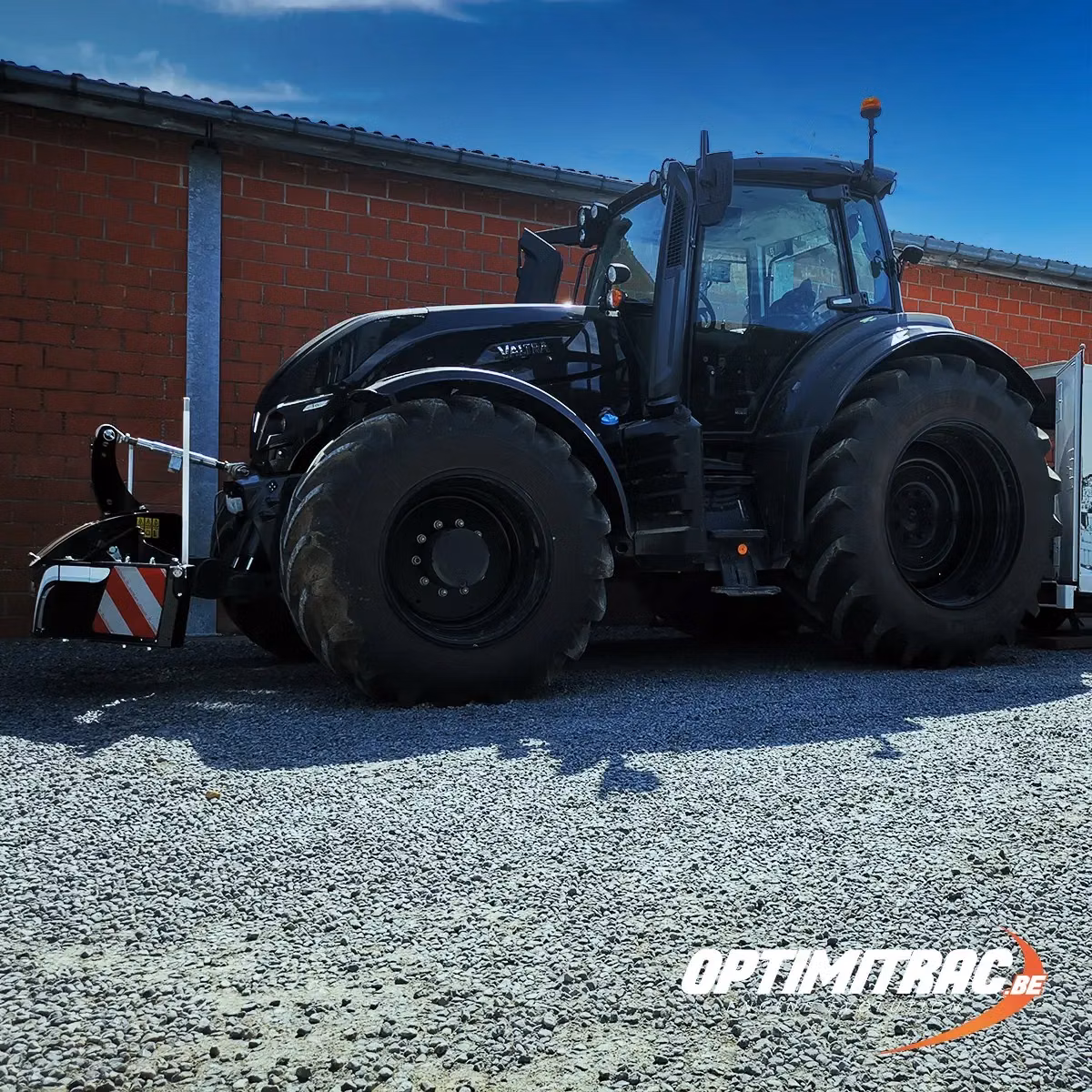Black tractor with large tires fitted with a front weight bumper, parked on gravel beside a brick building