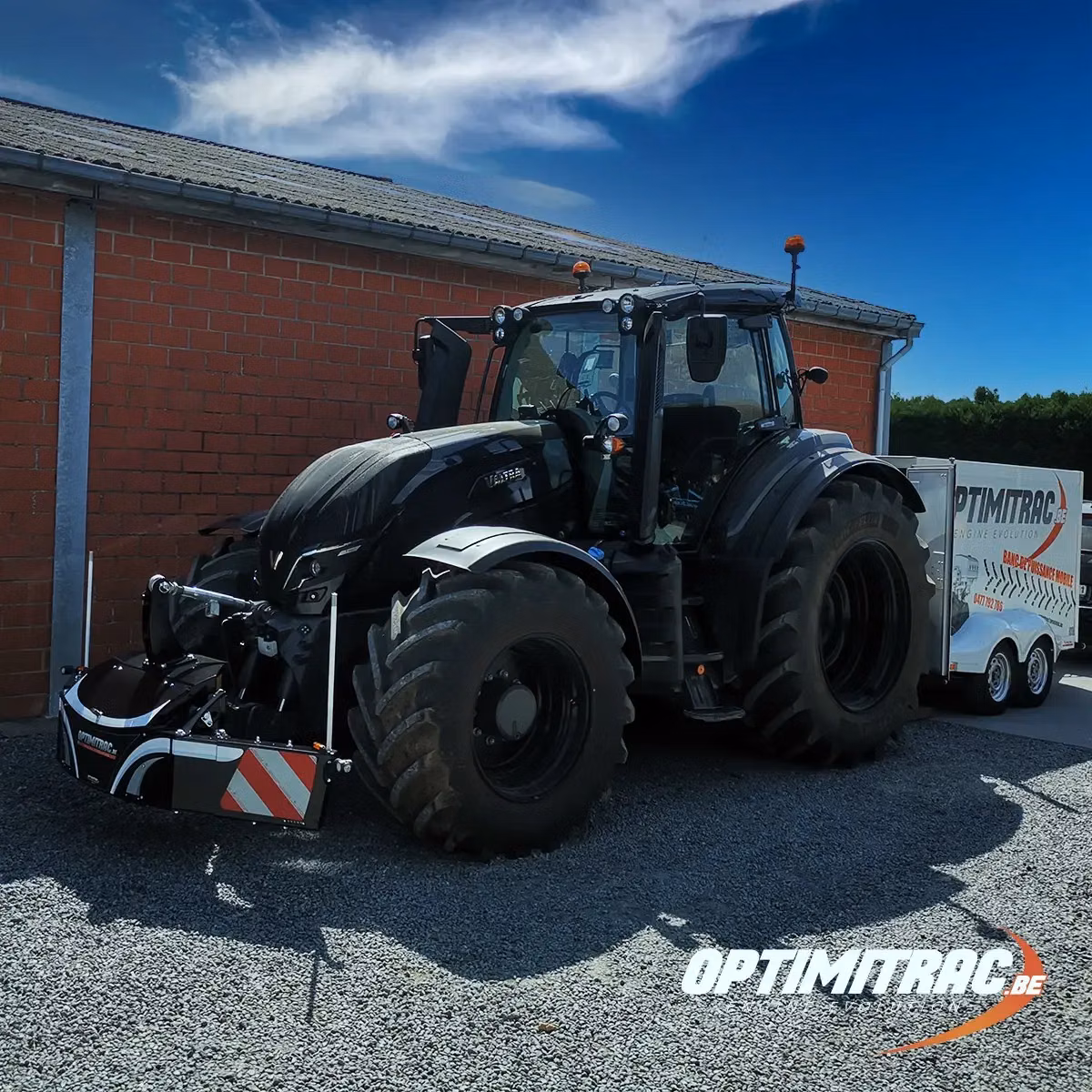 Black tractor with a front weight bumper attachment parked on gravel beside a brick building, trailer in background