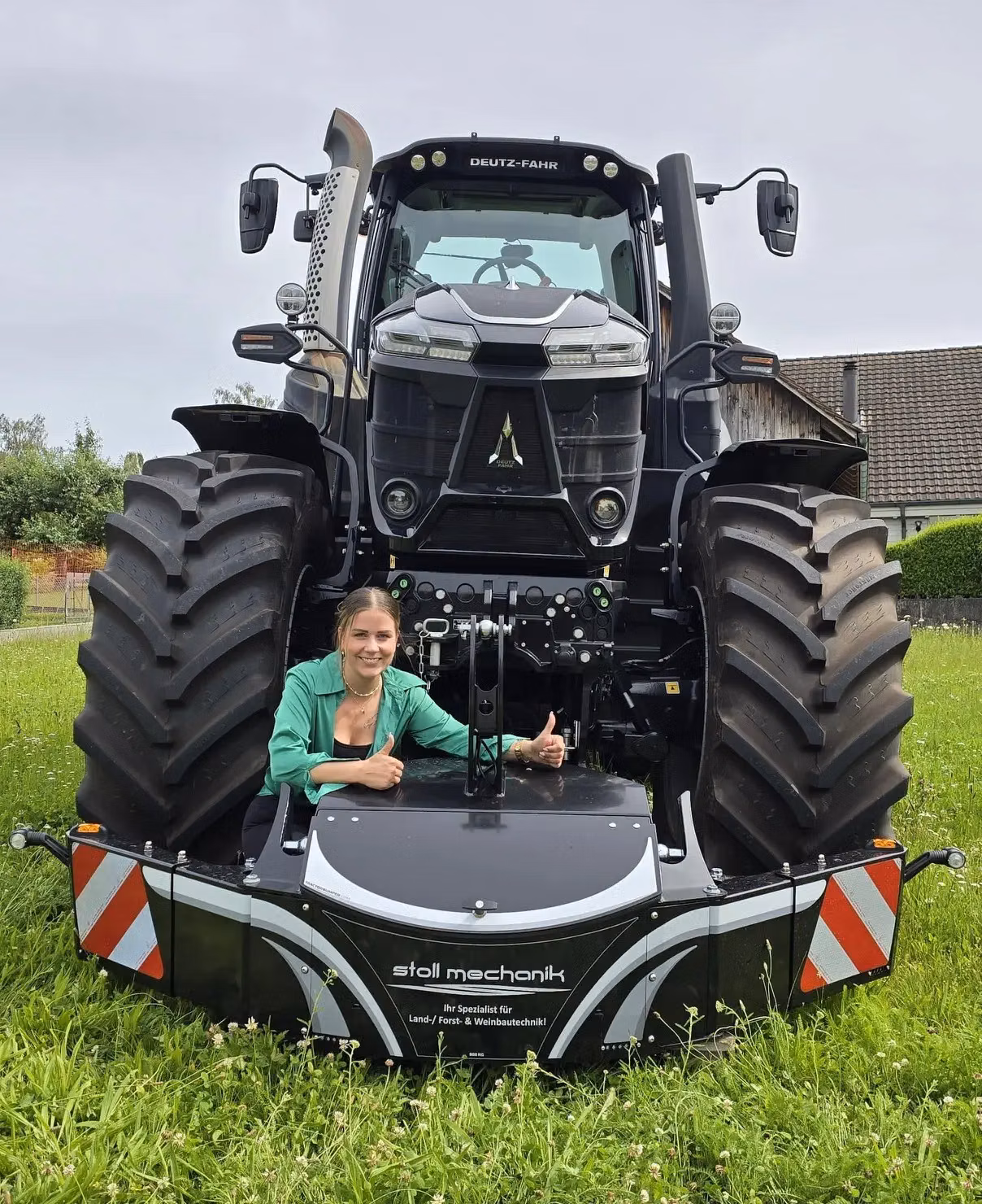 A woman sits in front of a large tractor equipped with a black front weight bumper on grass giving thumbs up