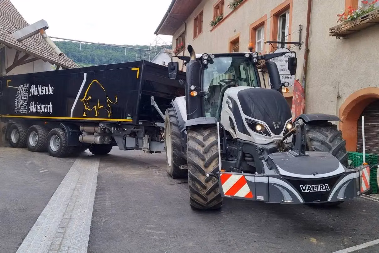 A Valtra tractor with a front weight bumper featuring safety markings and LED lights is towing a large black trailer