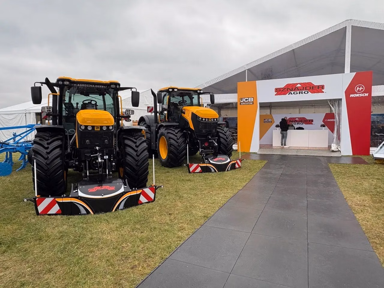 Two yellow tractors with large front weight bumpers are displayed on grass near a white and red exhibition booth