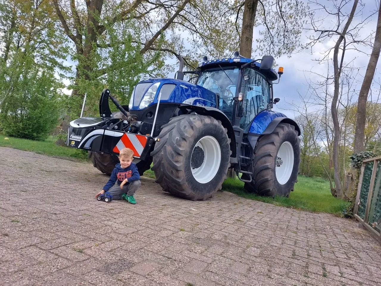 A large blue tractor with a front weight bumper featuring red and white stripes, parked on paving with a young child playing