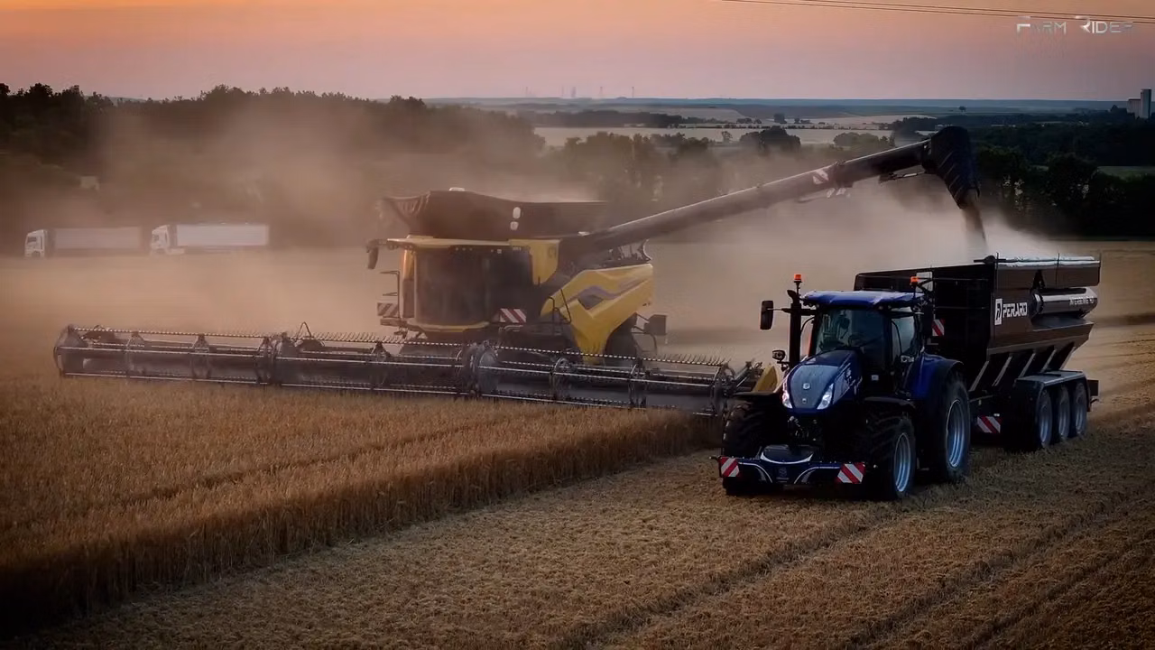 A blue tractor with a front weight bumper and attached trailer drives beside a yellow combine harvester in a dusty field