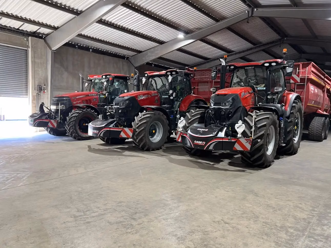 Three red tractors with front weight bumpers and trailers are parked indoors under a metal roof