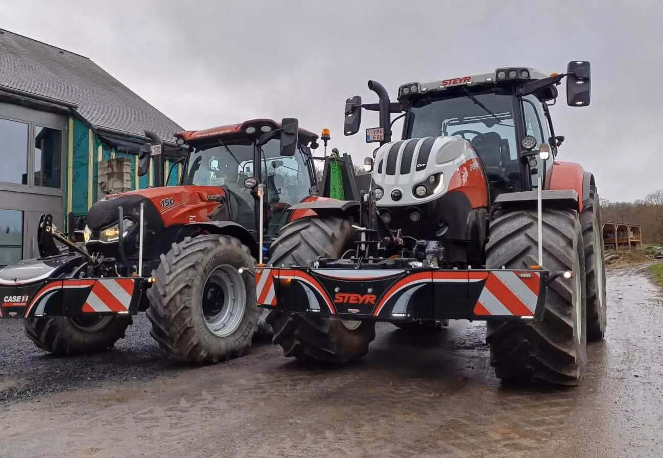 Two red tractors with large front weight bumpers featuring reflective stripes are parked on a farm driveway