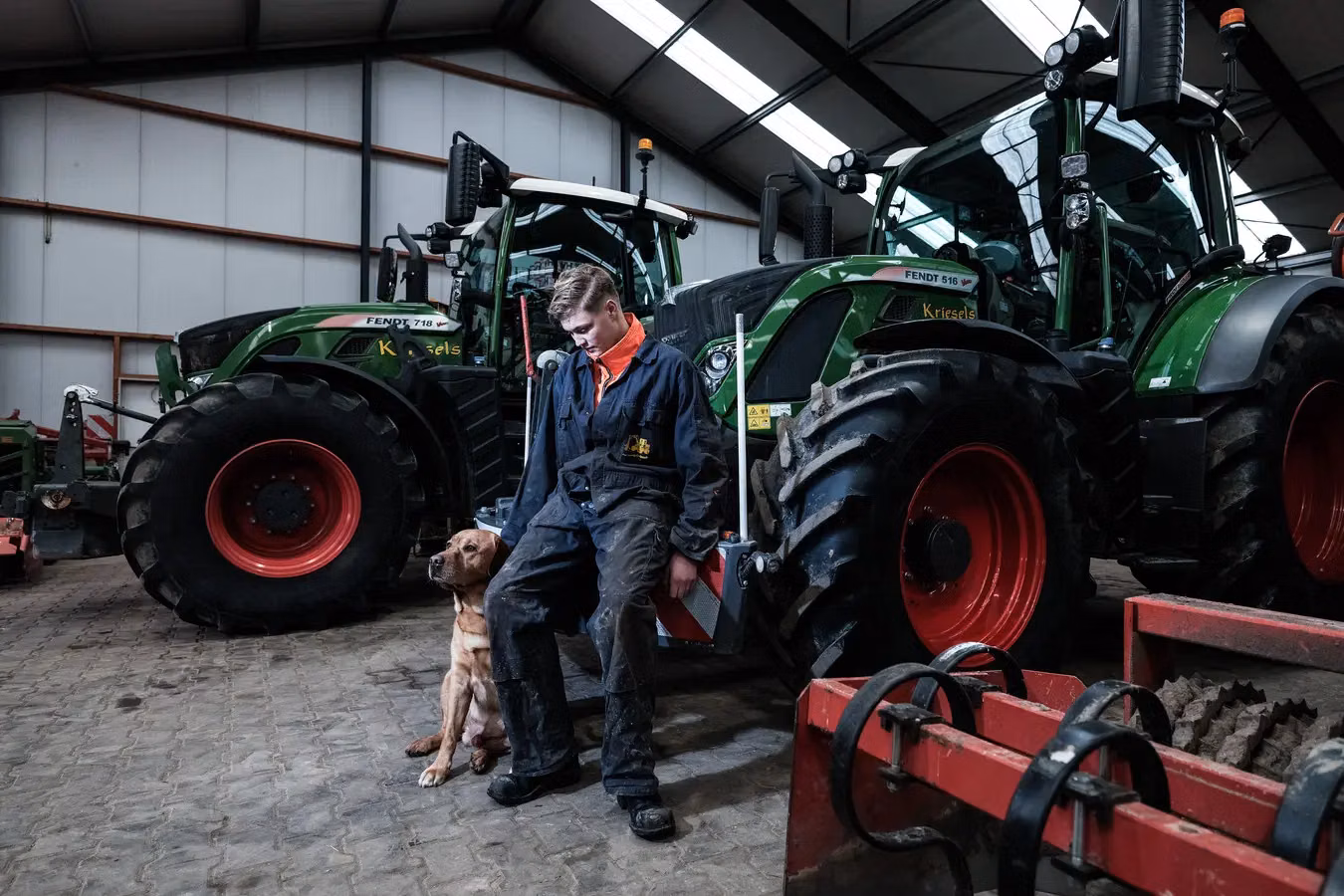 A person in work clothes sits on a front weight bumper attached to a green tractor beside a brown dog in a shed