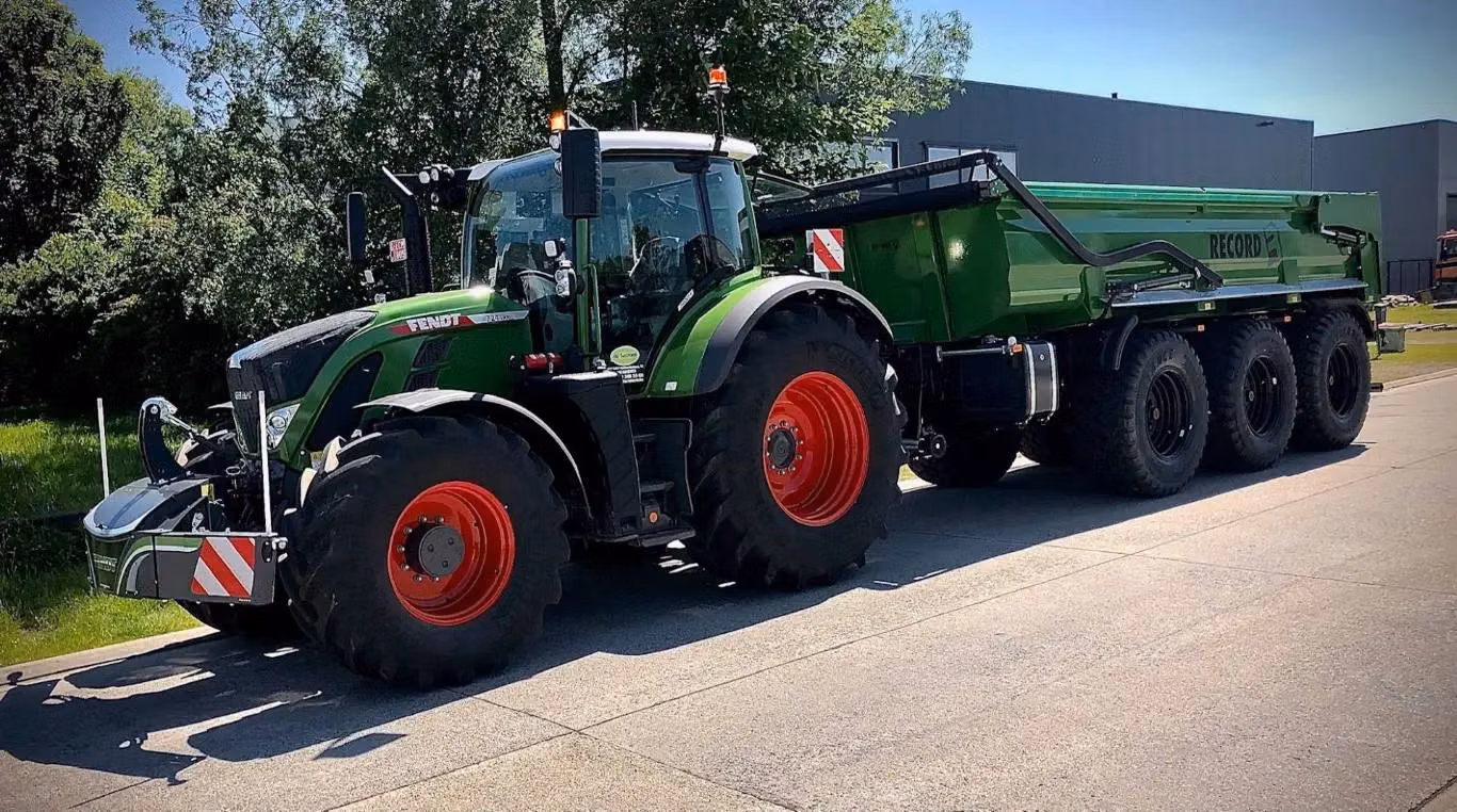 A green tractor with a front weight bumper attached, towing a large green trailer with six wheels on a sunny day