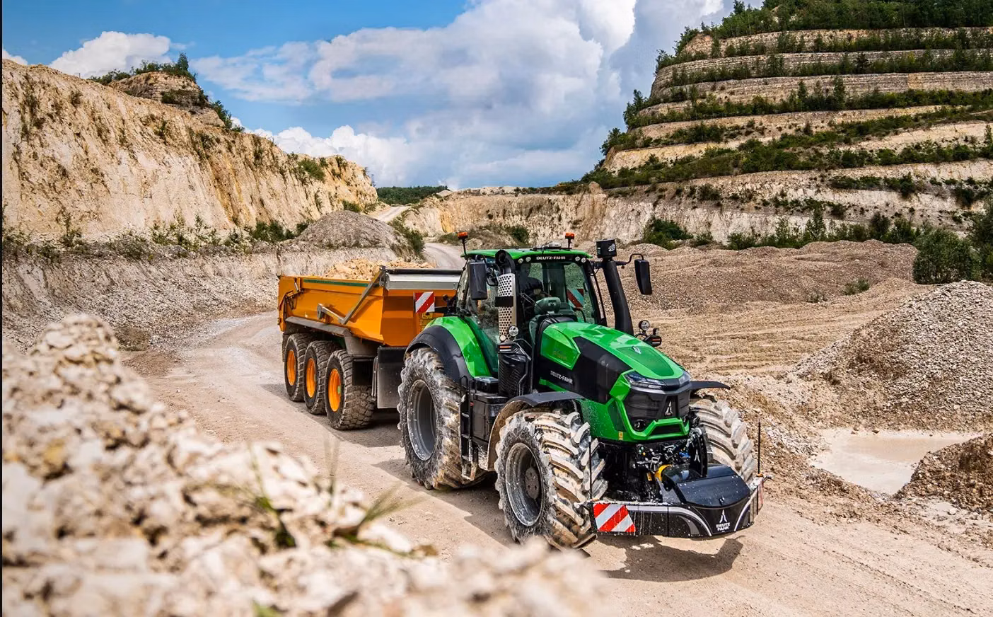 Green tractor with a front weight bumper and an orange trailer driving on a dirt road in a rocky quarry
