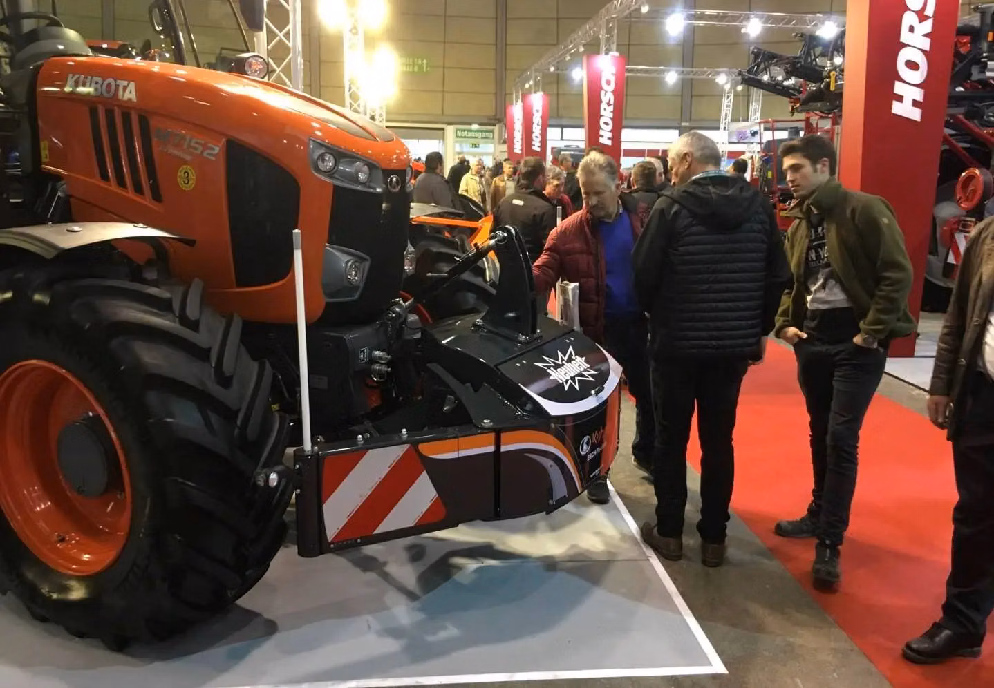 A group of people examine a tractor equipped with a front weight bumper and safety markings at an indoor expo
