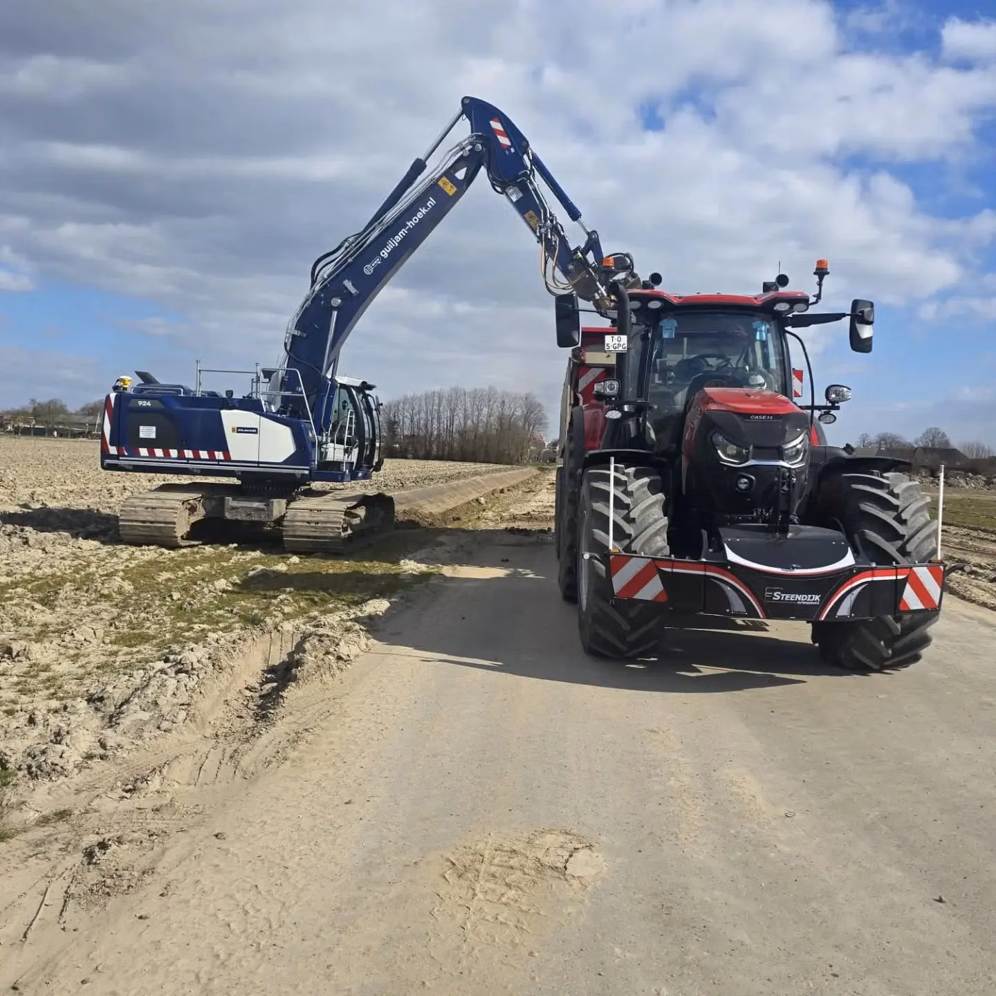 A red tractor with a front weight bumper is parked on a rural road next to a blue excavator on tracks
