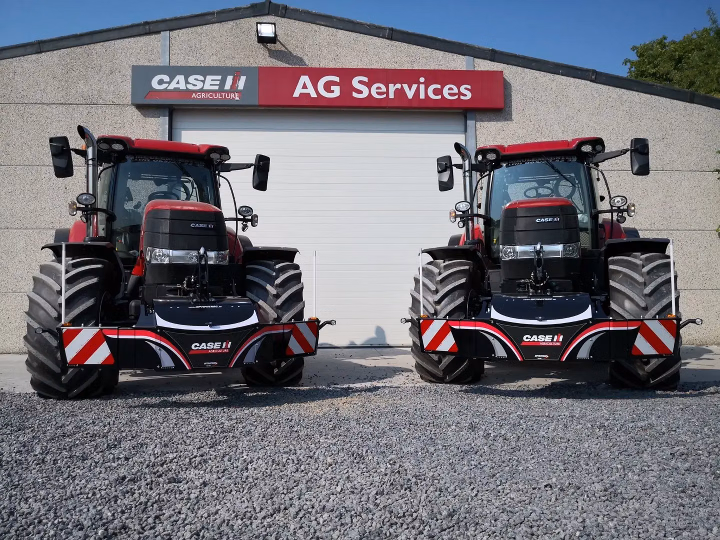 Two red tractors with large front weight bumpers are parked on gravel in front of an AG Services building