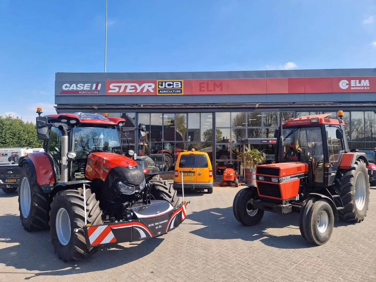 Two red tractors parked outside a dealership, with the left tractor fitted with a large front weight bumper