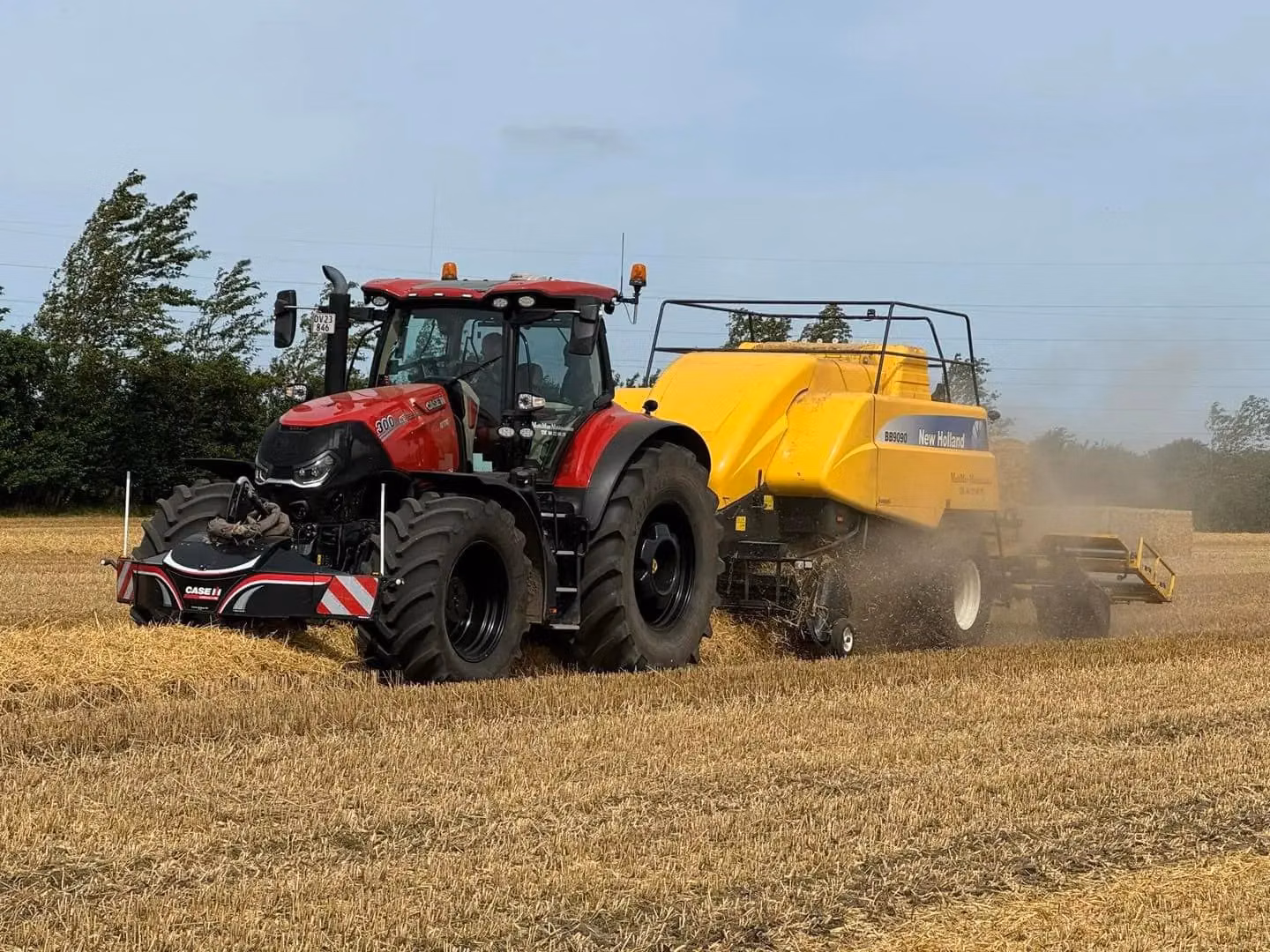 A red tractor with a front weight bumper attached is pulling a large yellow baler across a harvested field