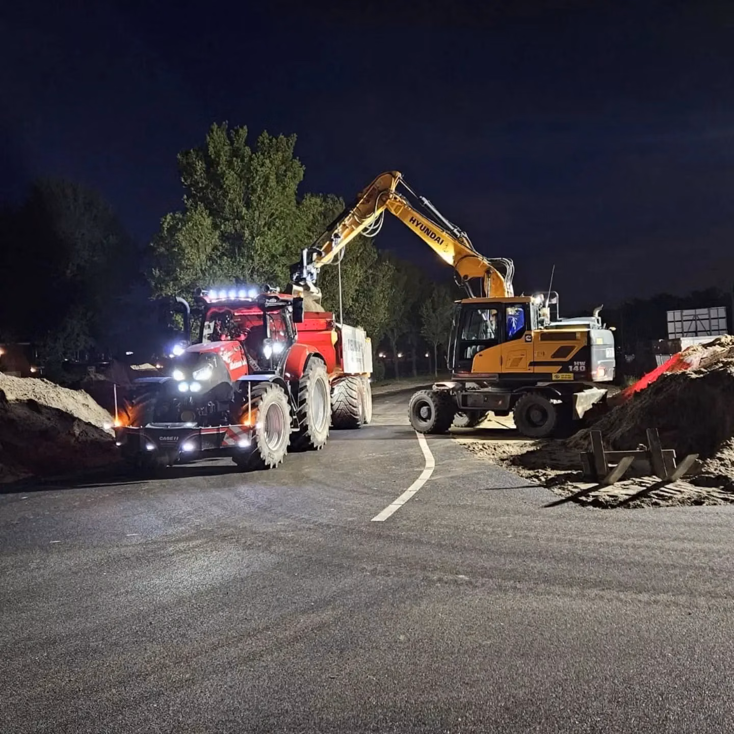A red tractor with a front weight bumper is parked on a road at night next to an excavator loading dirt.