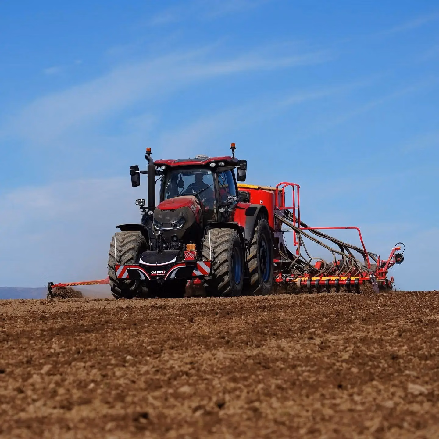 A red tractor with a front weight bumper and seeding equipment attached works in a ploughed field under blue sky