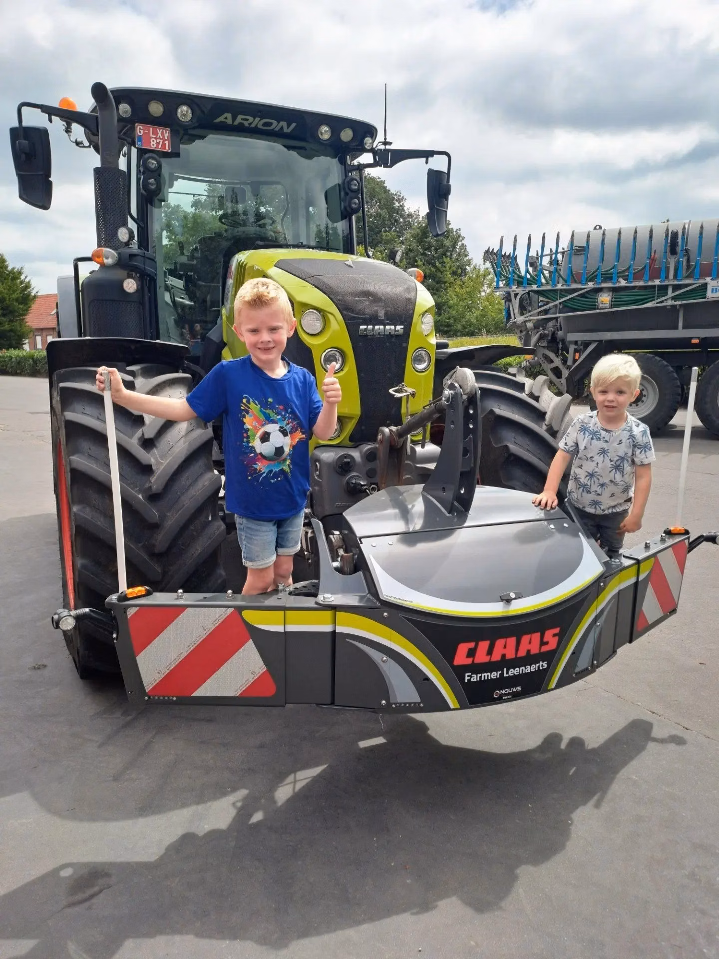 Two children stand on a CLAAS front weight bumper attached to a green tractor outdoors on a paved surface