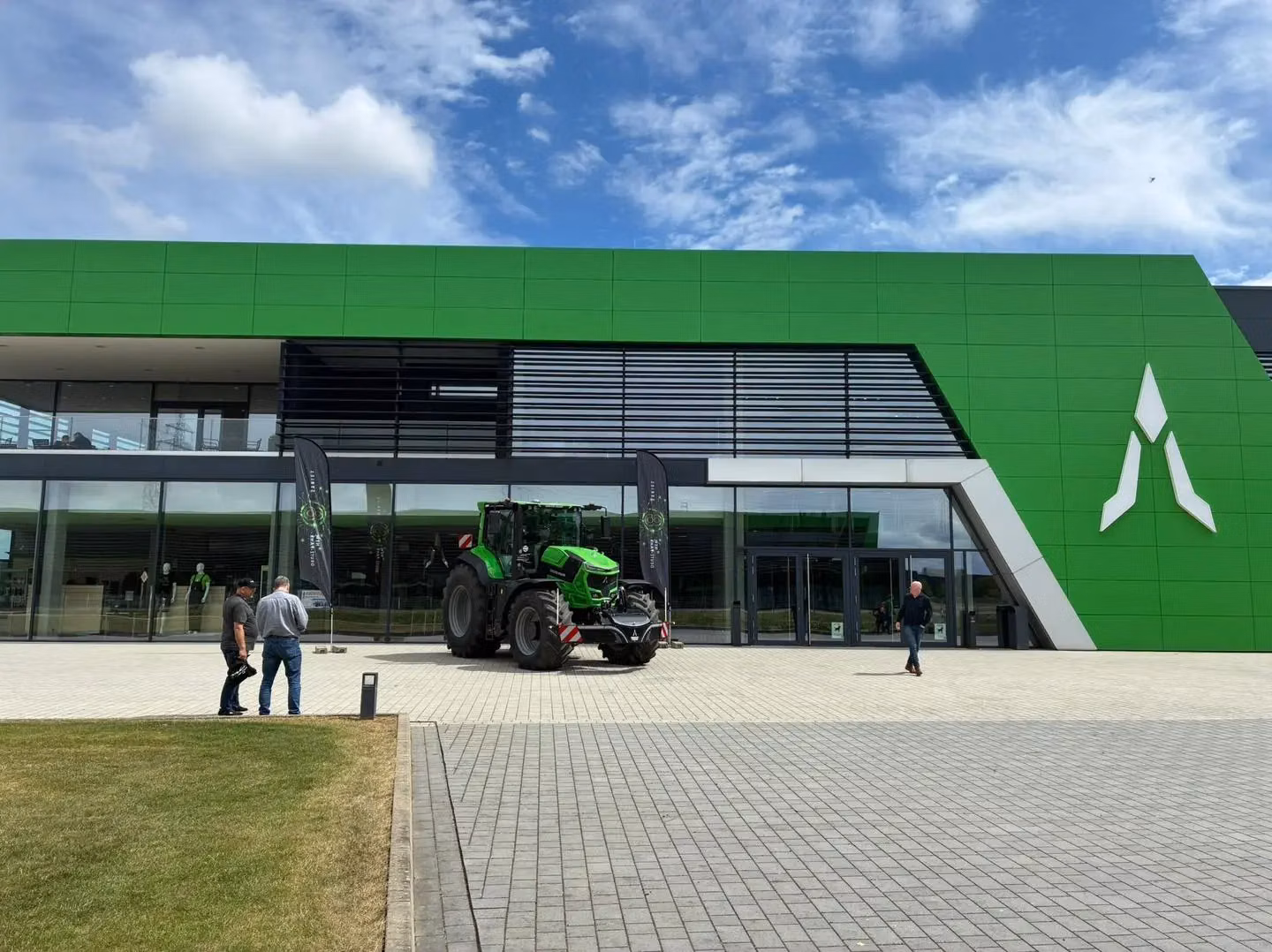 A green tractor with a front weight bumper is parked outside a modern building with people nearby.