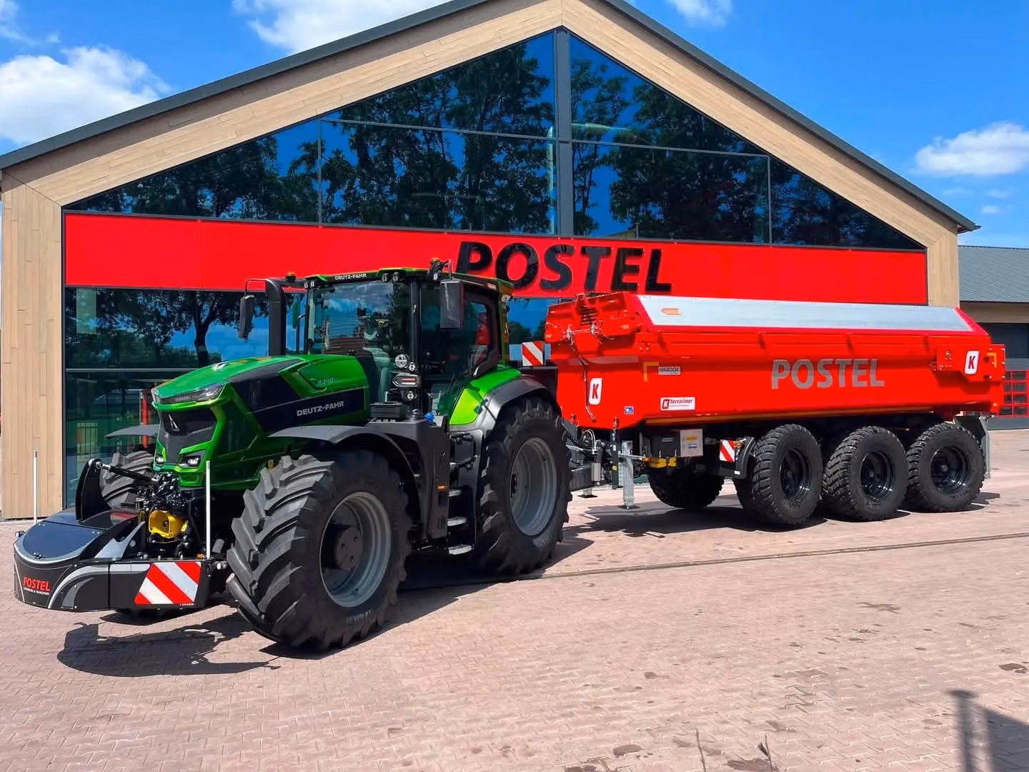Green tractor with a large front weight bumper attached, parked in front of a building with POSTEL signage