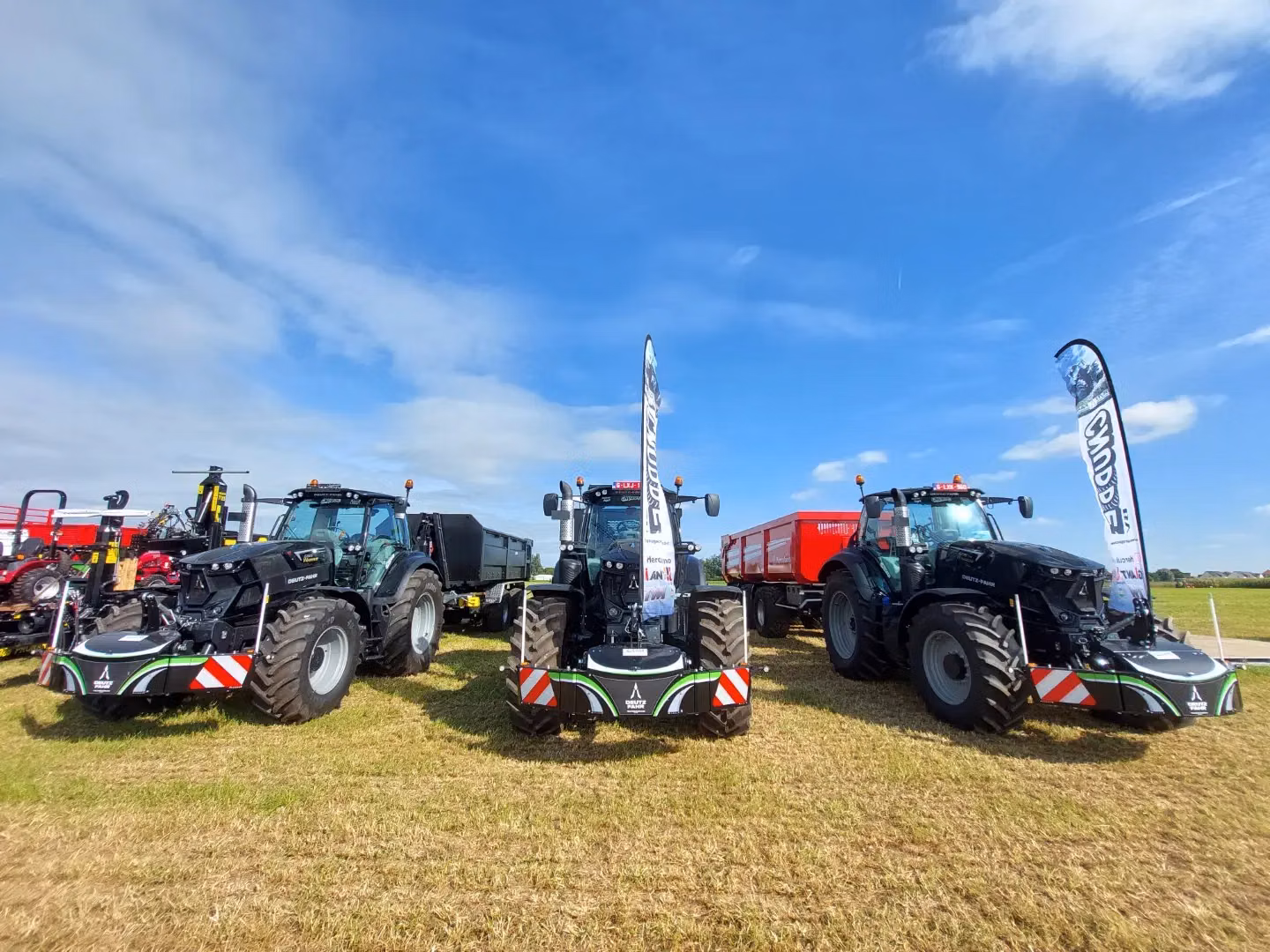Three large tractors fitted with front weight bumpers are displayed on grass under a sunny blue sky with flags nearby