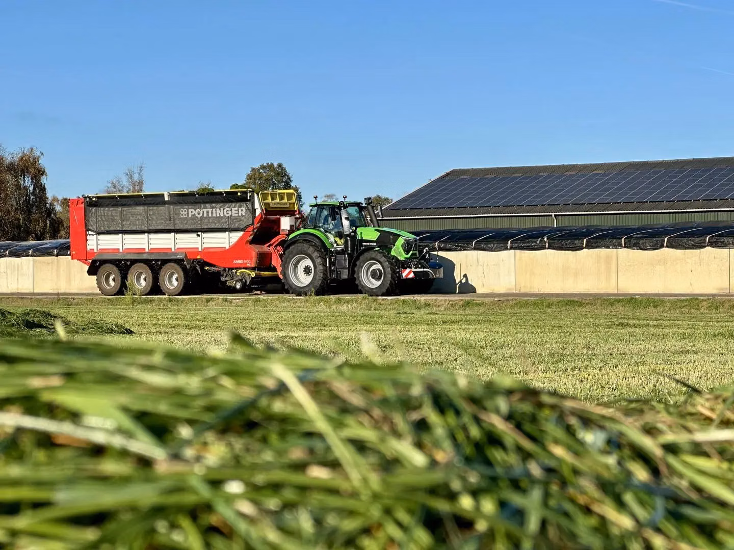 A green tractor with a front weight bumper is towing a large red trailer beside a silage pit on a sunny day