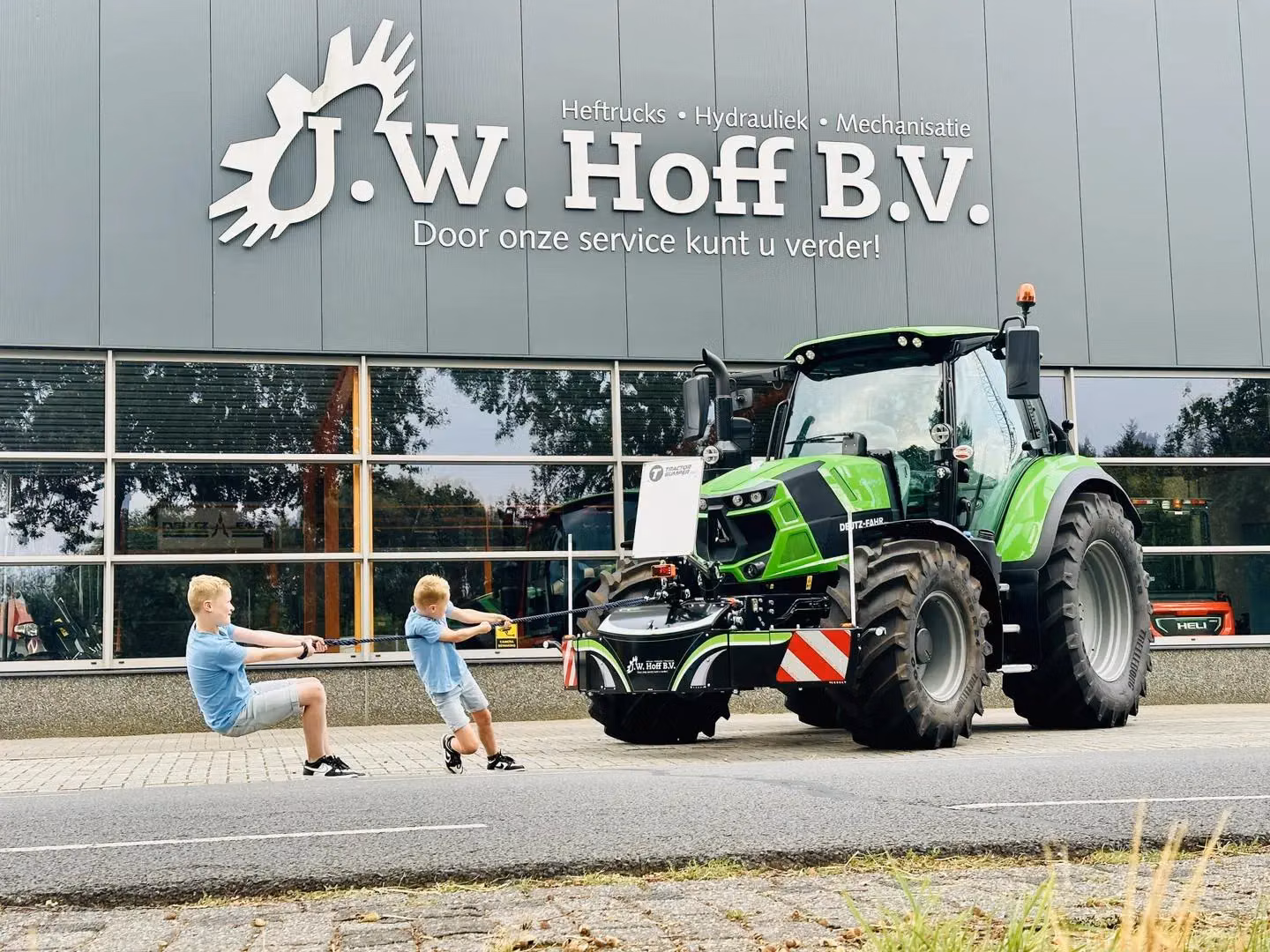 Two children pull on a rope attached to a green tractor with a front weight bumper outside a building