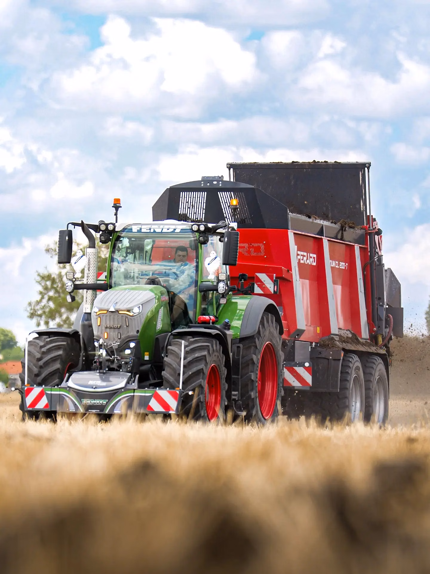 Green tractor with front weight bumper attached, pulling a large red trailer across a field under a cloudy sky