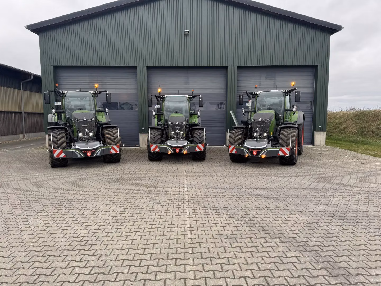 Three green tractors with front weight bumpers and LED lights are parked in front of a building with closed garage doors