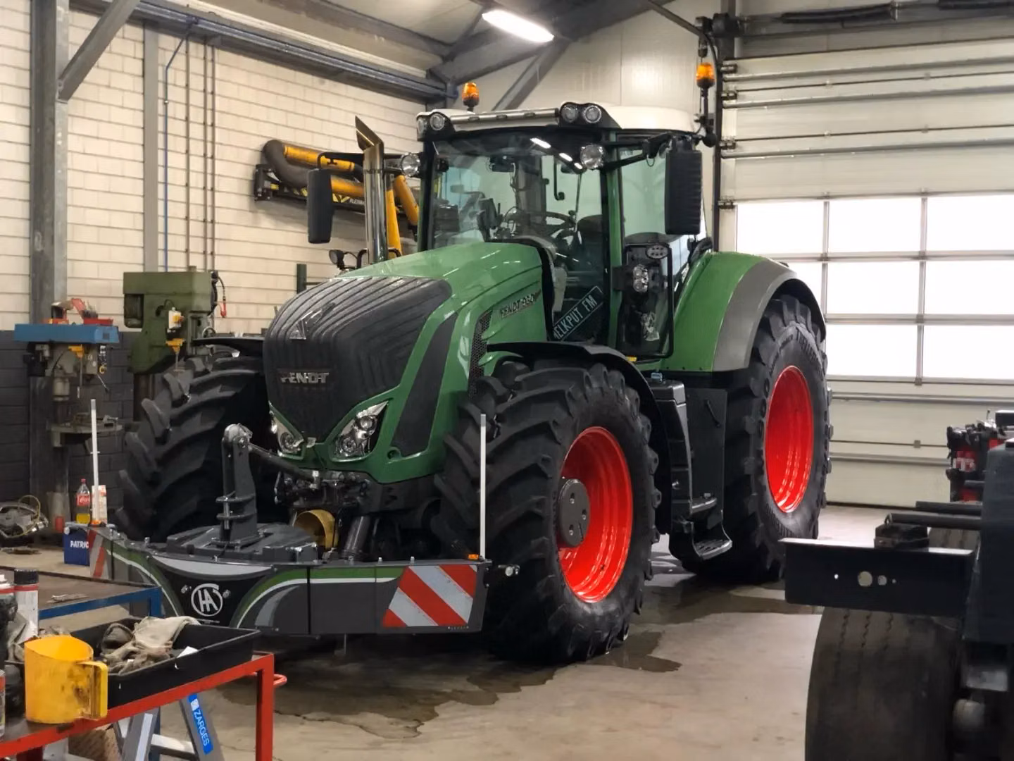A green tractor with a large front weight bumper is parked inside a workshop with tools and machinery nearby