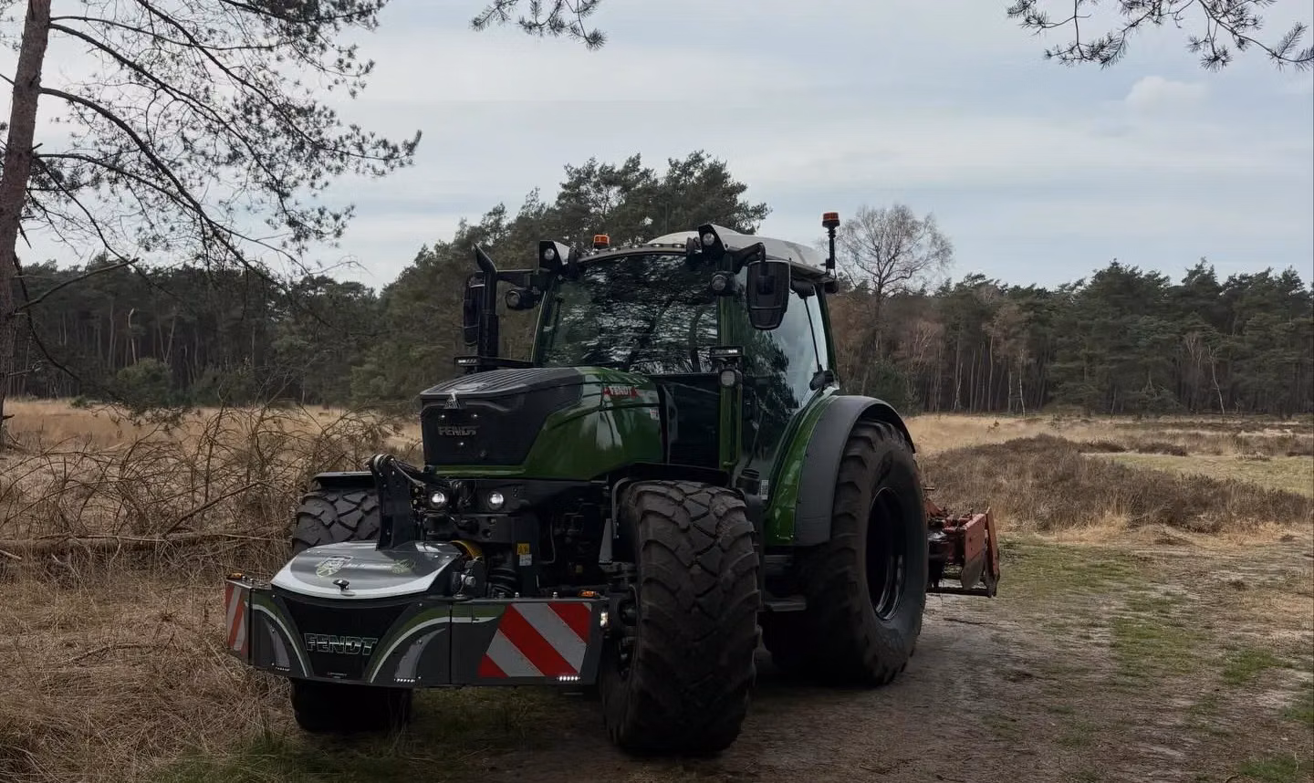 Green tractor with a front weight bumper attachment parked on a dirt path near a grassy field and forest
