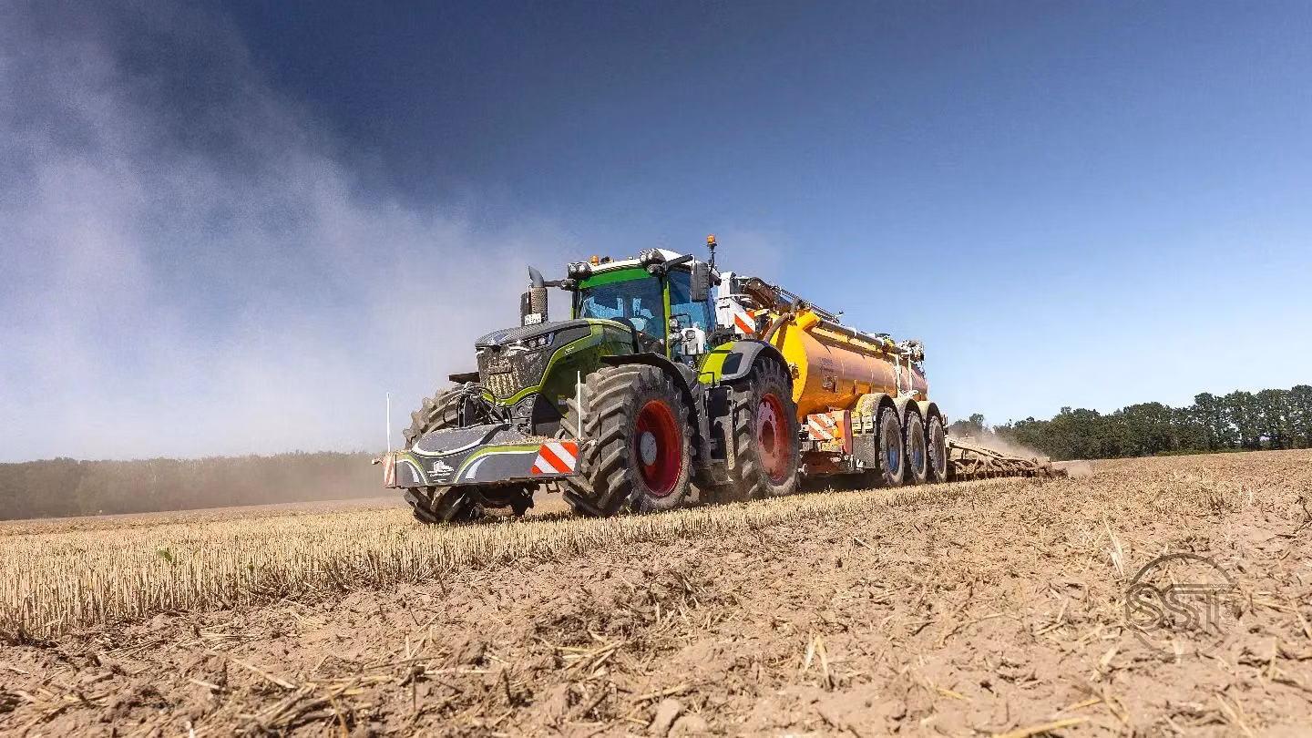 A tractor with a front weight bumper attached works in a dry field pulling a large yellow agricultural trailer