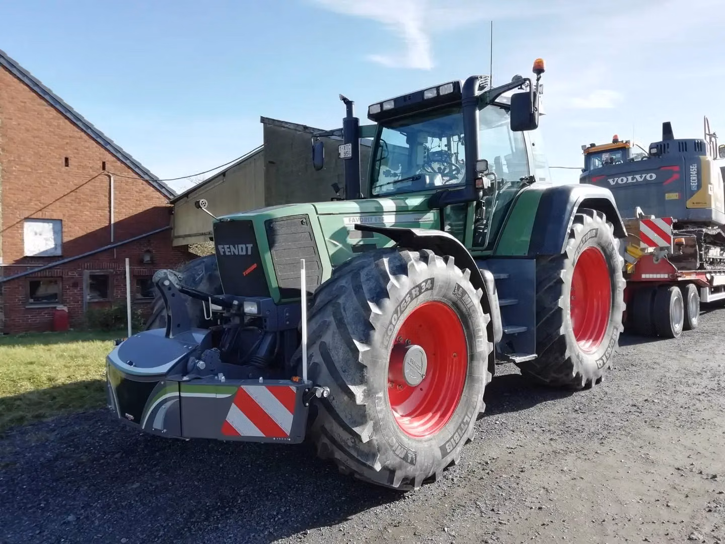 Green tractor with a front weight bumper attached, parked on gravel near buildings and a trailer in the background