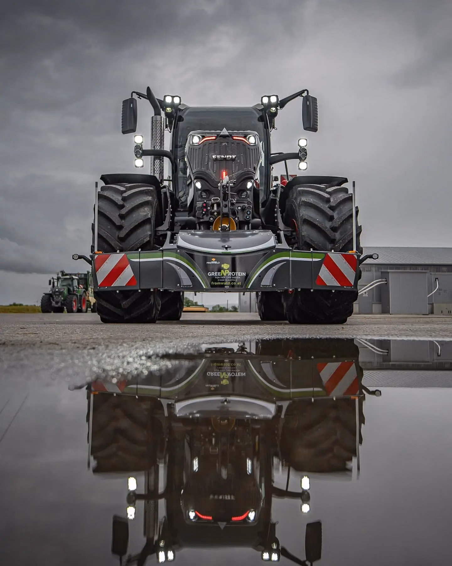 Large tractor with mounted front weight bumper featuring LED lights, reflected in a water puddle on an overcast day