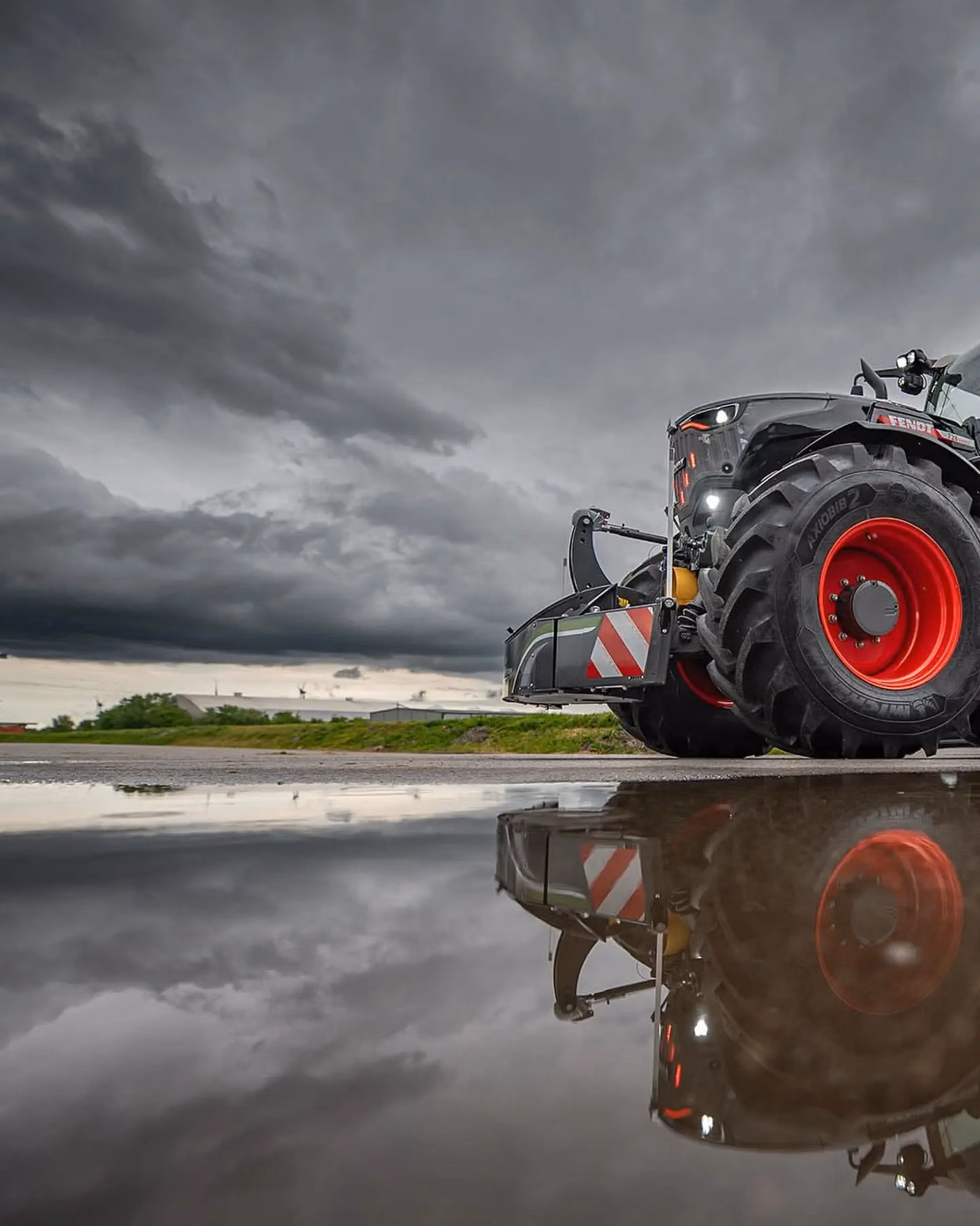 Large tractor with a front weight bumper and LED lights reflected in a puddle under a dramatic cloudy sky