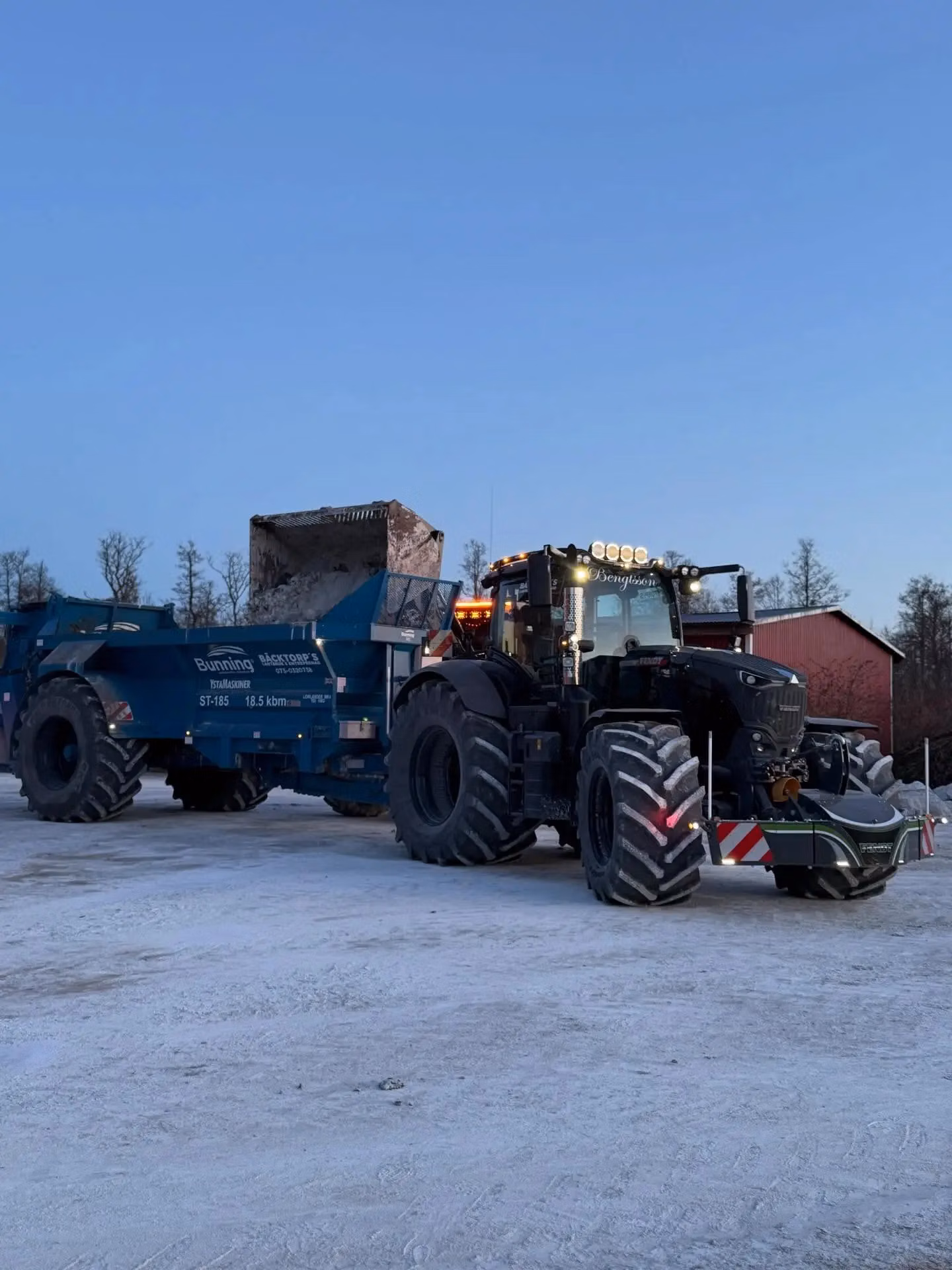 A tractor with a front weight bumper and warning stripes is parked on a snowy surface, attached to a large blue trailer
