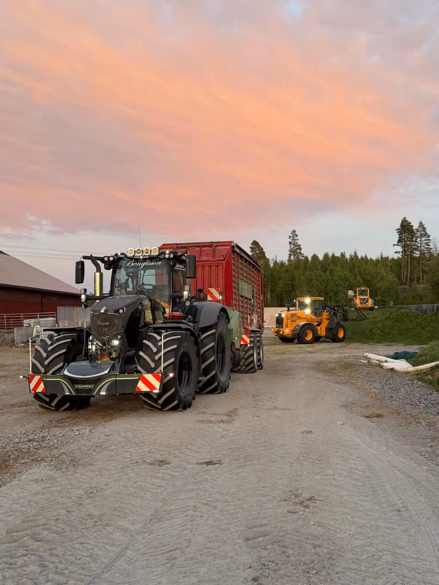 Large tractor with dual front wheels and a front weight bumper, attached to a red trailer on a farm at sunset