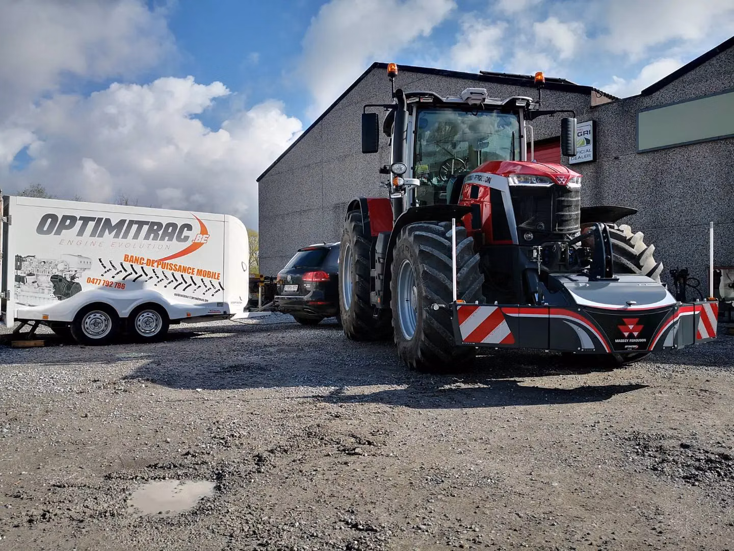 Red and black tractor with a Massey Ferguson front weight bumper parked on gravel near a trailer and a car