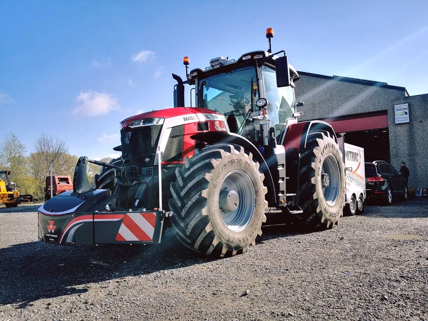 A red tractor fitted with a front weight bumper is parked outside a building on a gravel surface.