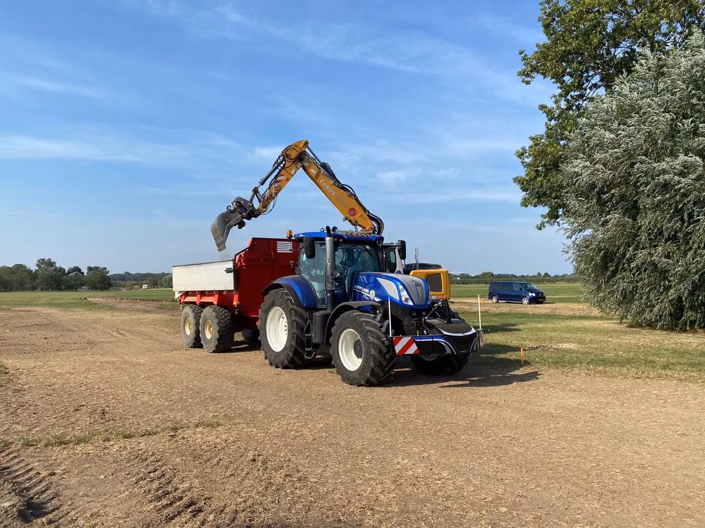 A blue tractor with a front weight bumper attached is pulling a red trailer being loaded by a yellow excavator arm