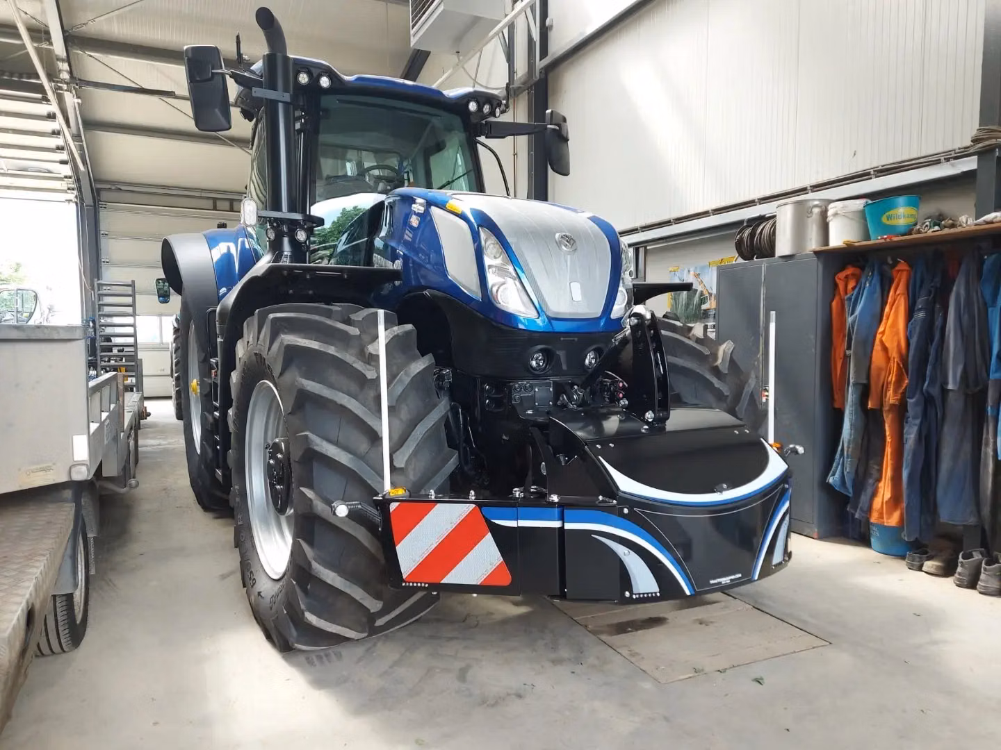 Blue tractor indoors fitted with a large black front weight bumper featuring reflective red and white stripes