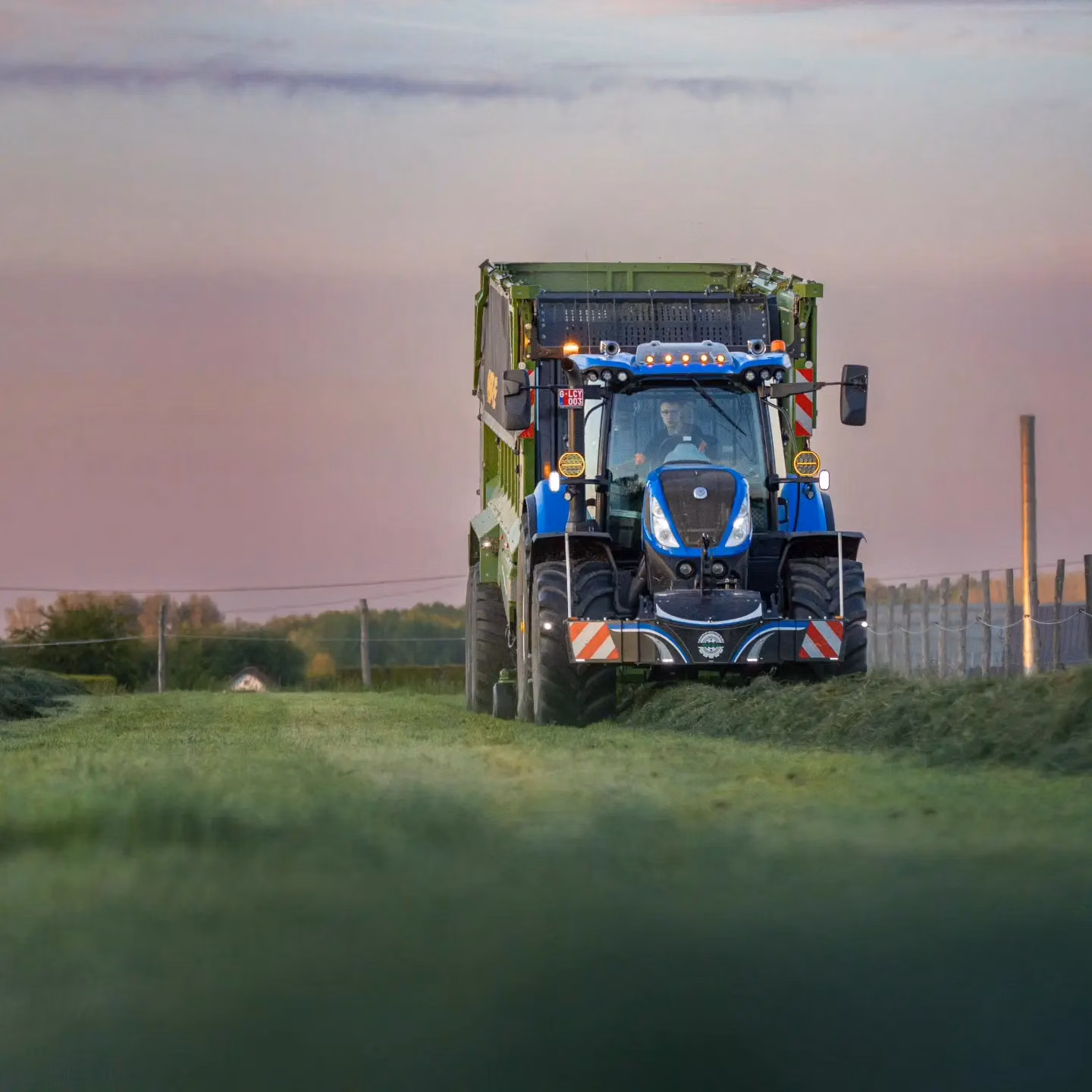 Blue tractor with a front weight bumper and LED lights drives on a grassy path at sunset with a large green trailer