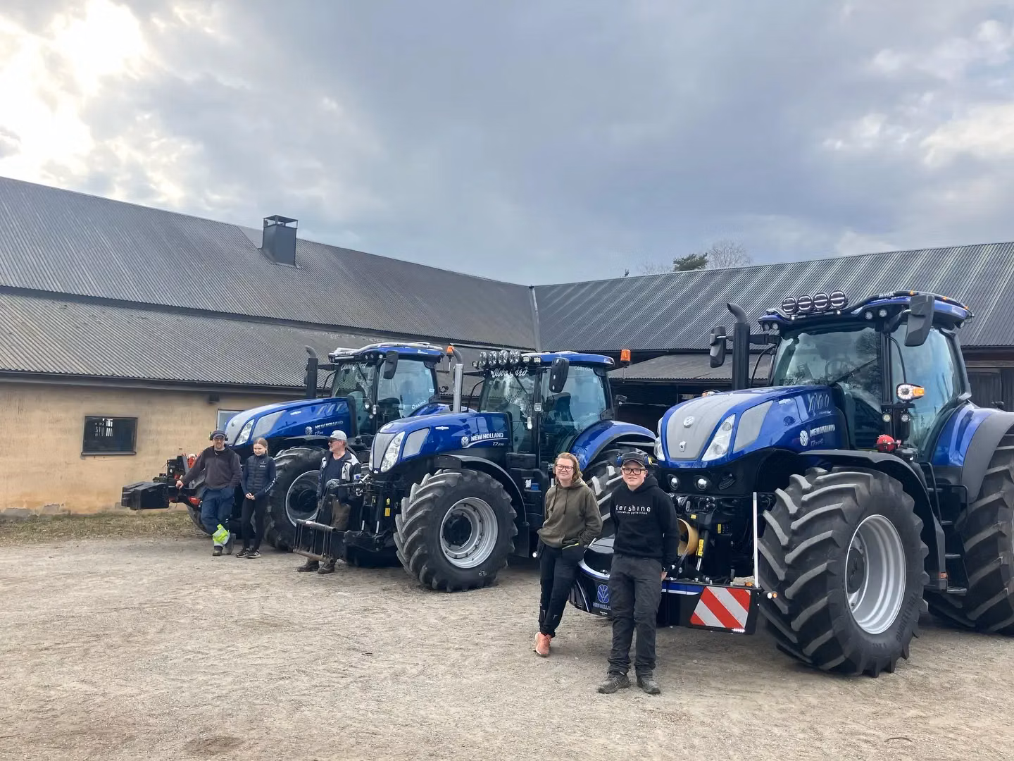 Five people stand in front of three large blue tractors with front weight bumpers outside a farm building