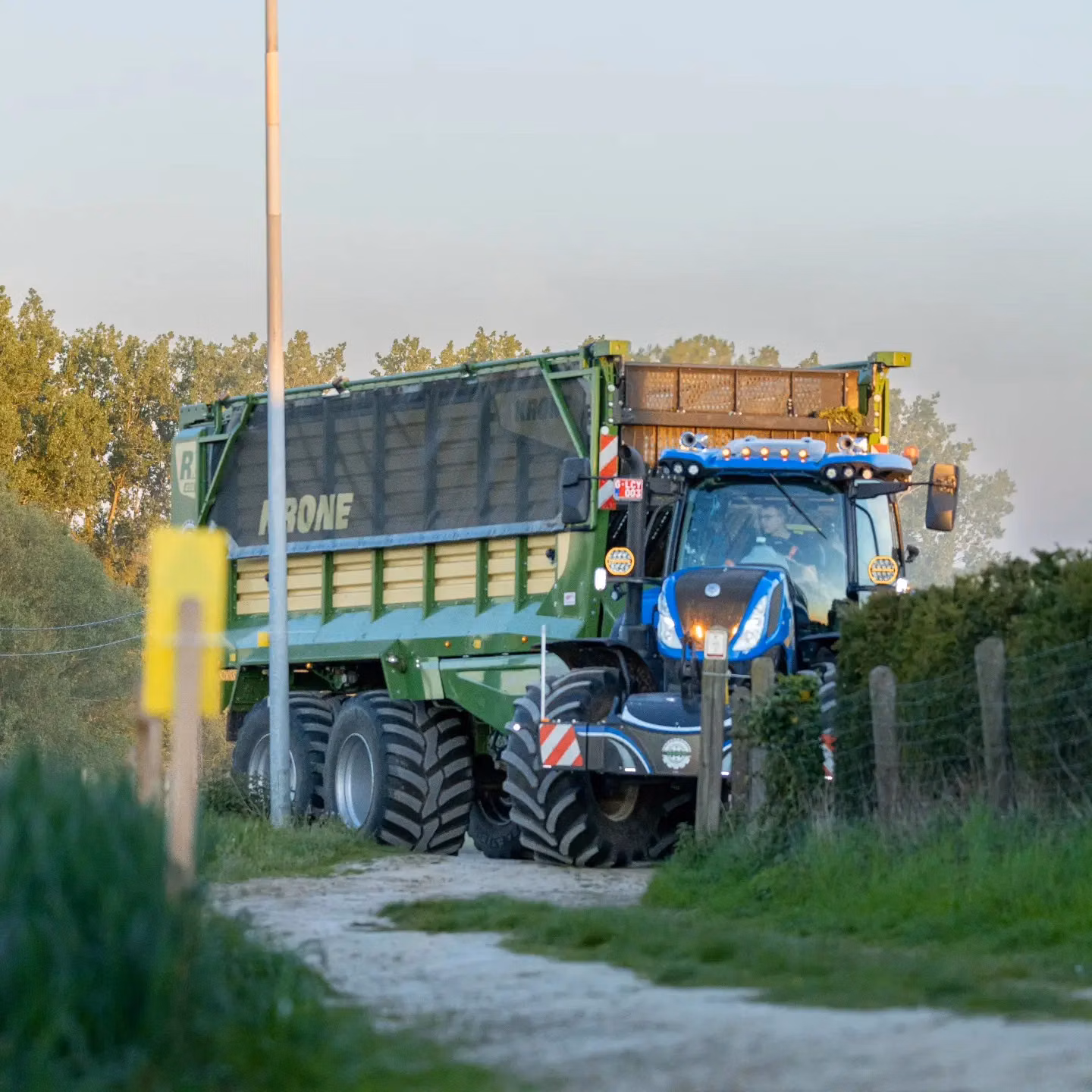 A blue tractor with a front weight bumper and LED lights pulls a large green trailer on a dirt track