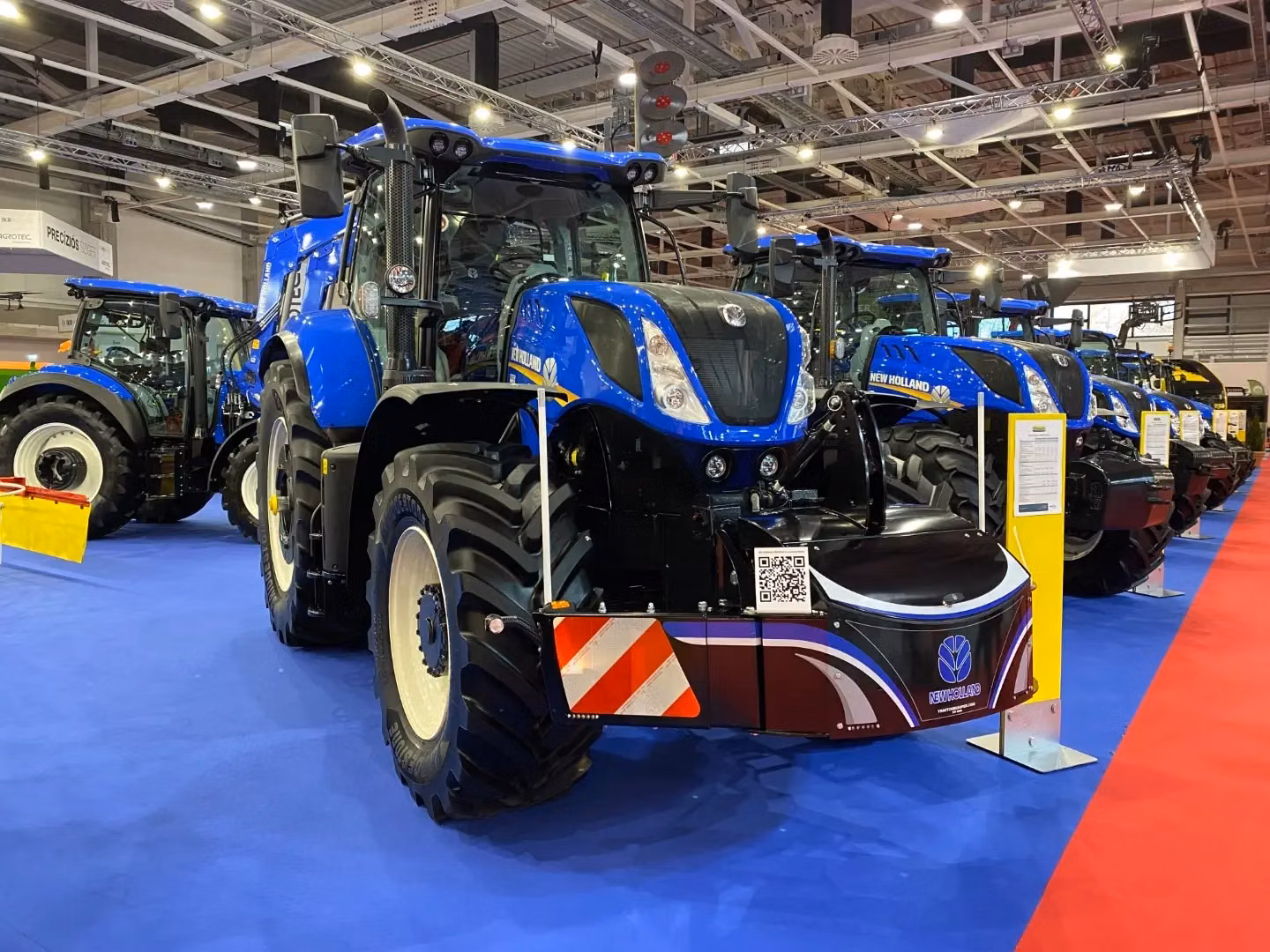 A row of blue tractors with large front weight bumpers on display indoors at an exhibition