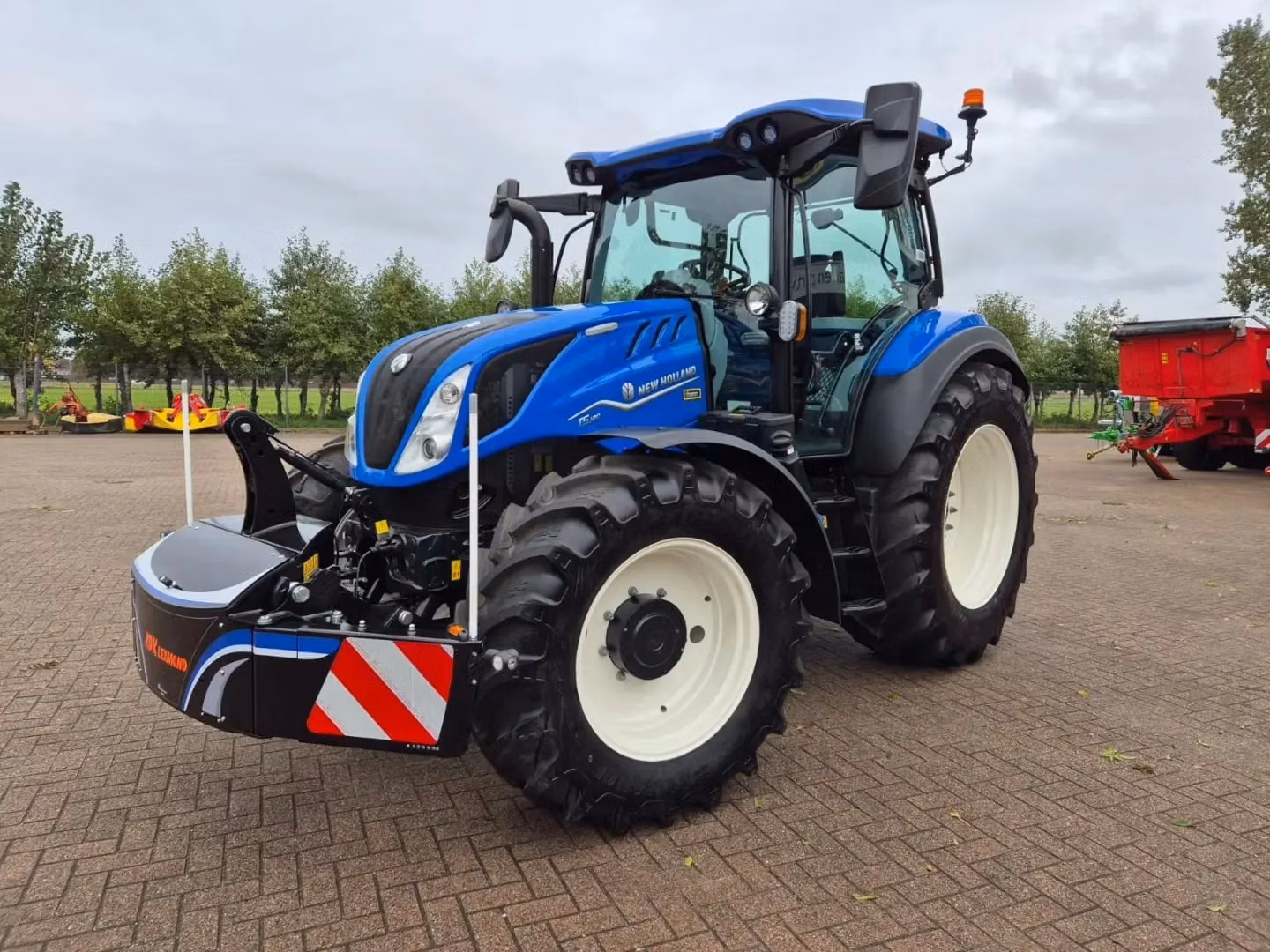 Blue tractor fitted with a front weight bumper featuring reflective red and white stripes, parked on paving