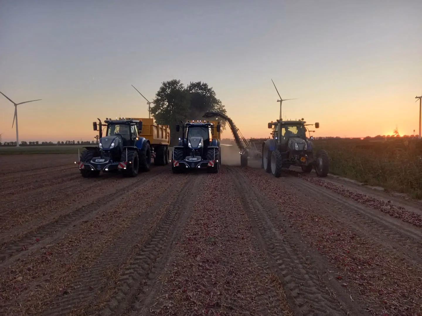Three blue tractors with front weight bumpers work in a field at sunset with wind turbines in the background