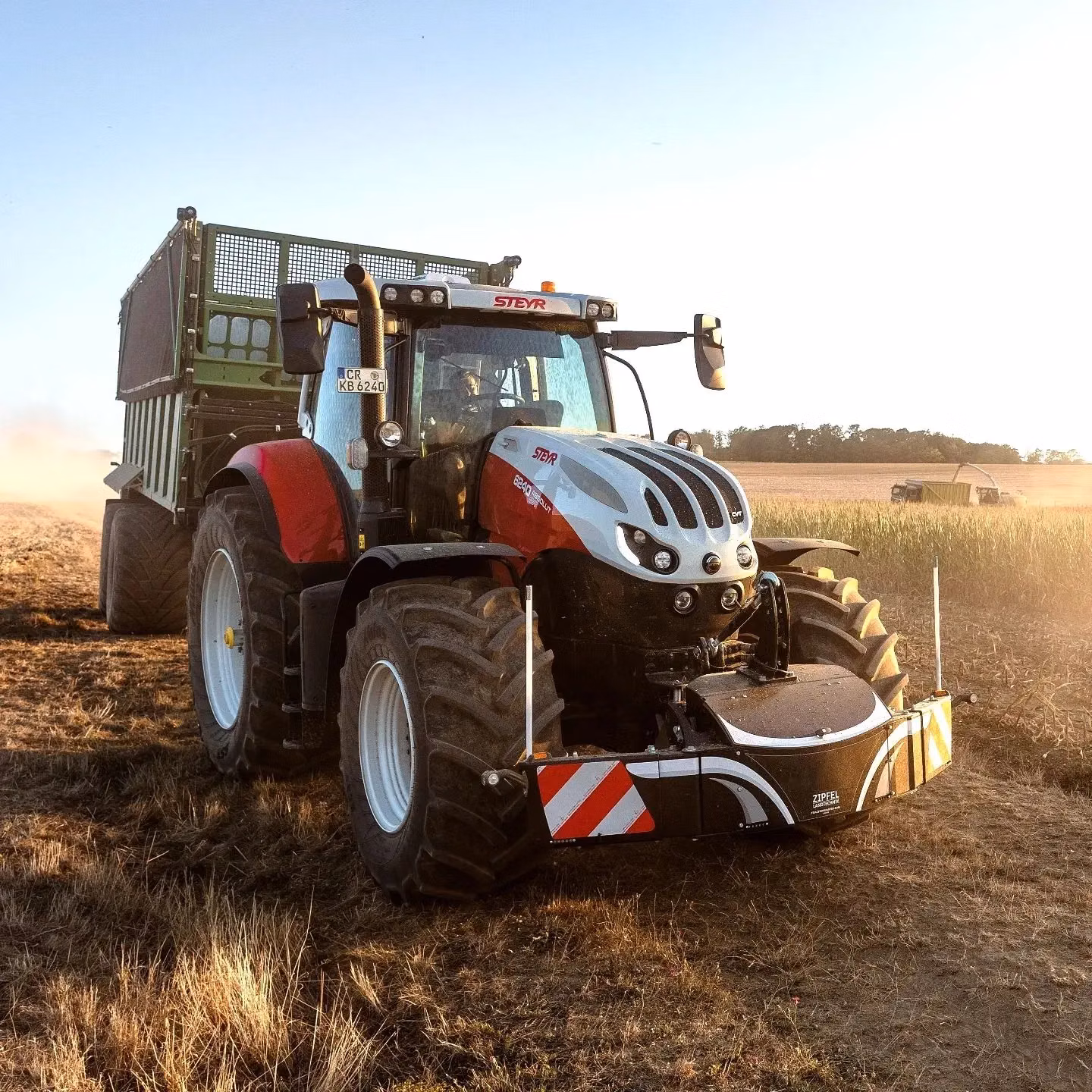 A tractor with a front weight bumper attached drives through a dry field with a trailer hitched behind it